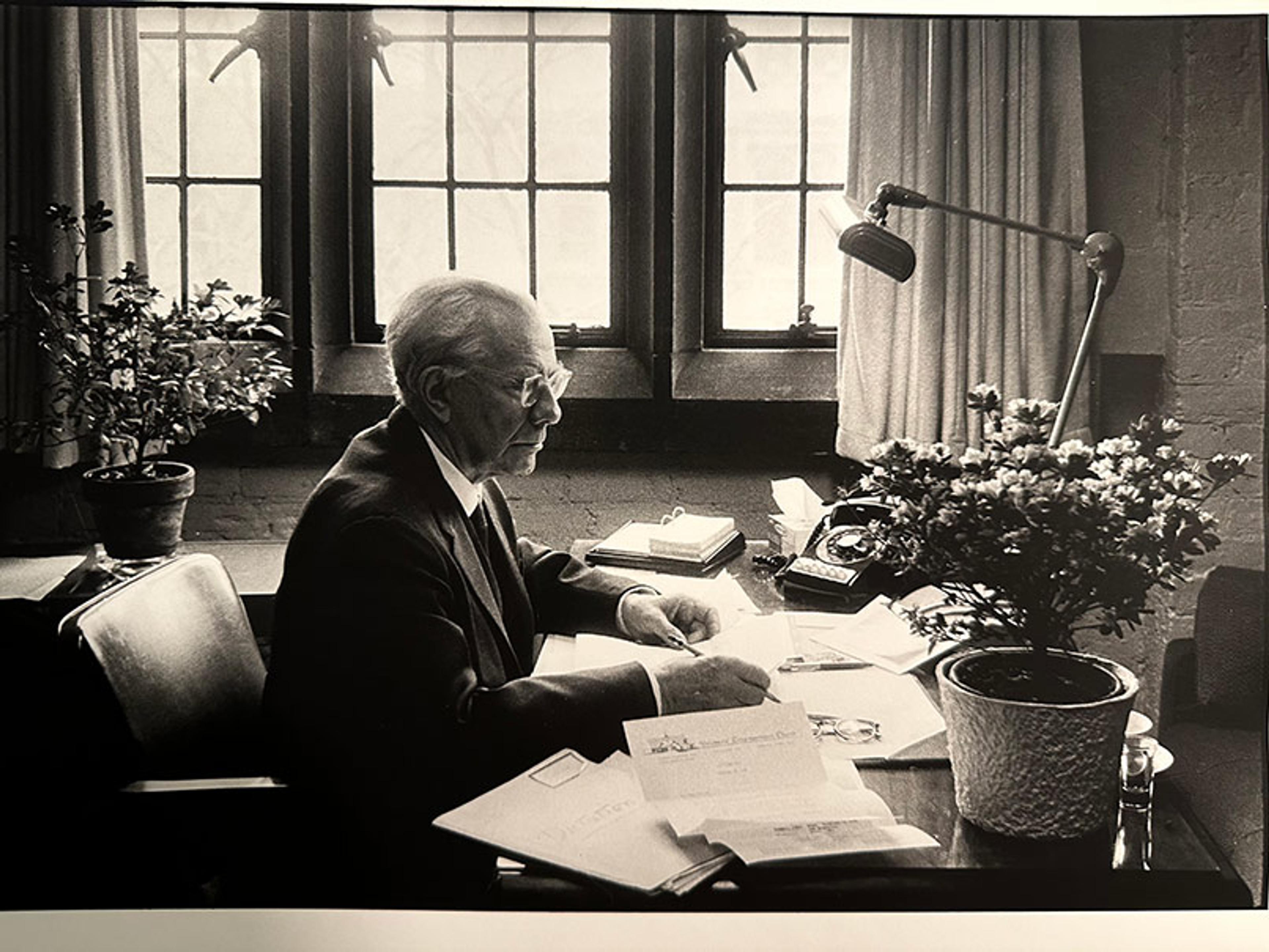 Black and white image of an elderly man in a suit, sitting at a desk covered with papers, beside a potted plant. He is writing or reading a document. Behind him, large paned windows let in light. A desk lamp, telephone, and additional paperwork are also visible on the desk.