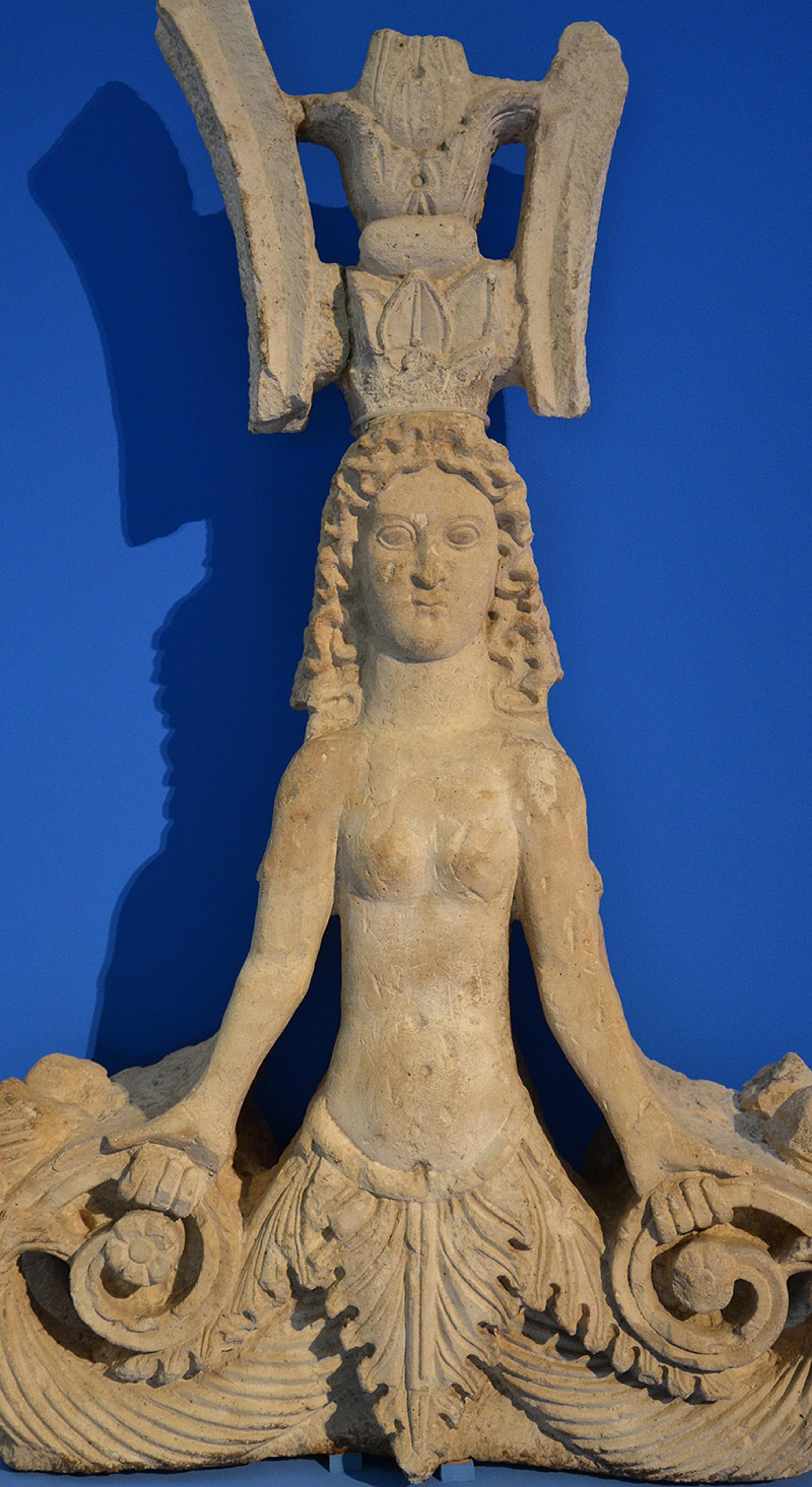 A snake goddess statue in stone. She is bare chested, faces forward and wears an elaborate headdress