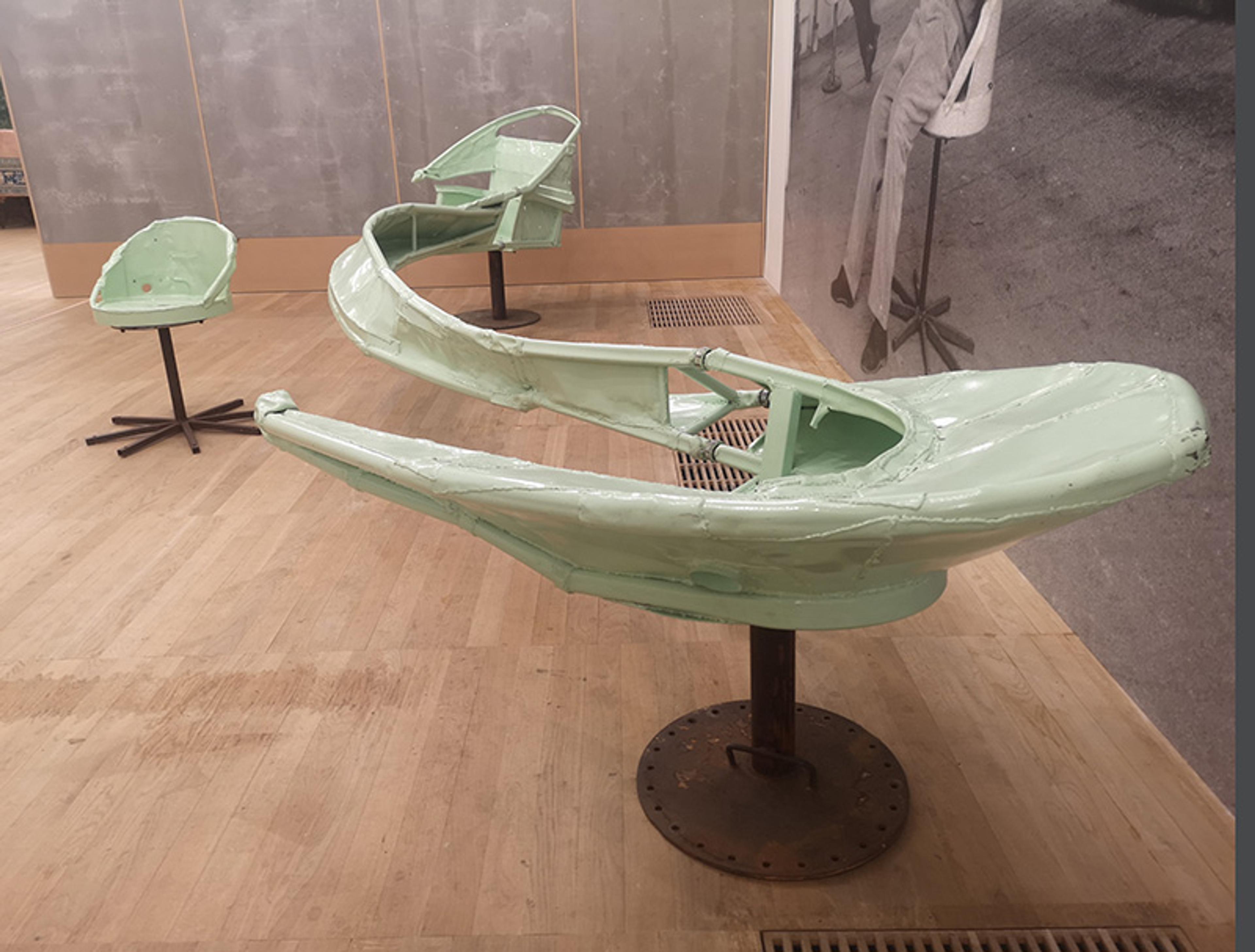 Three green, industrial metal structures mounted on stands are displayed on a wooden floor. The structures resemble abstract, aerodynamic forms. In the background, there is a monochrome mural of a person.