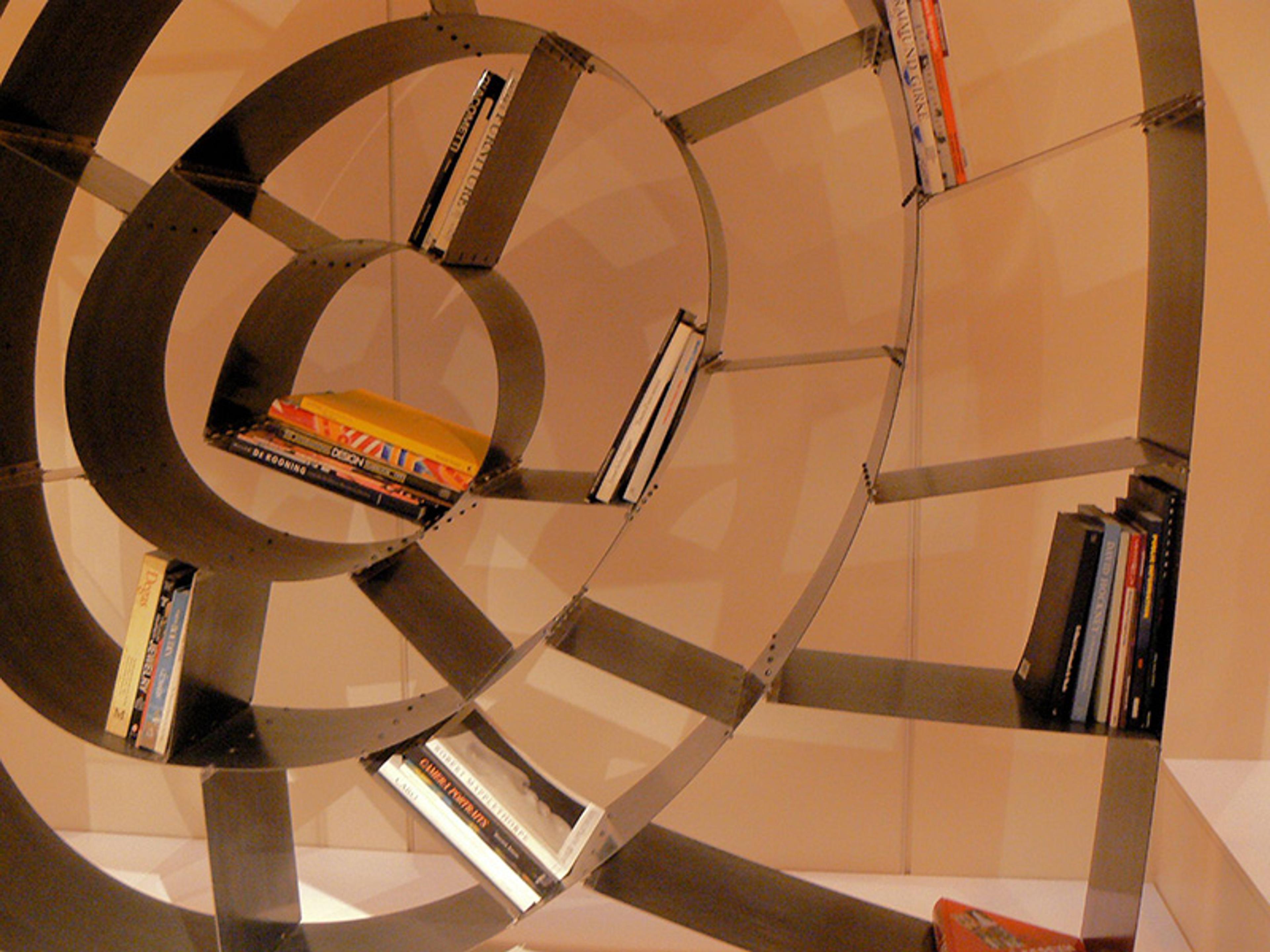 A unique bookshelf with a spiral design, holding various books on its shelves. The shelves emanate outward from the centre and are made of metal, contrasting against a light-coloured wall in the background.
