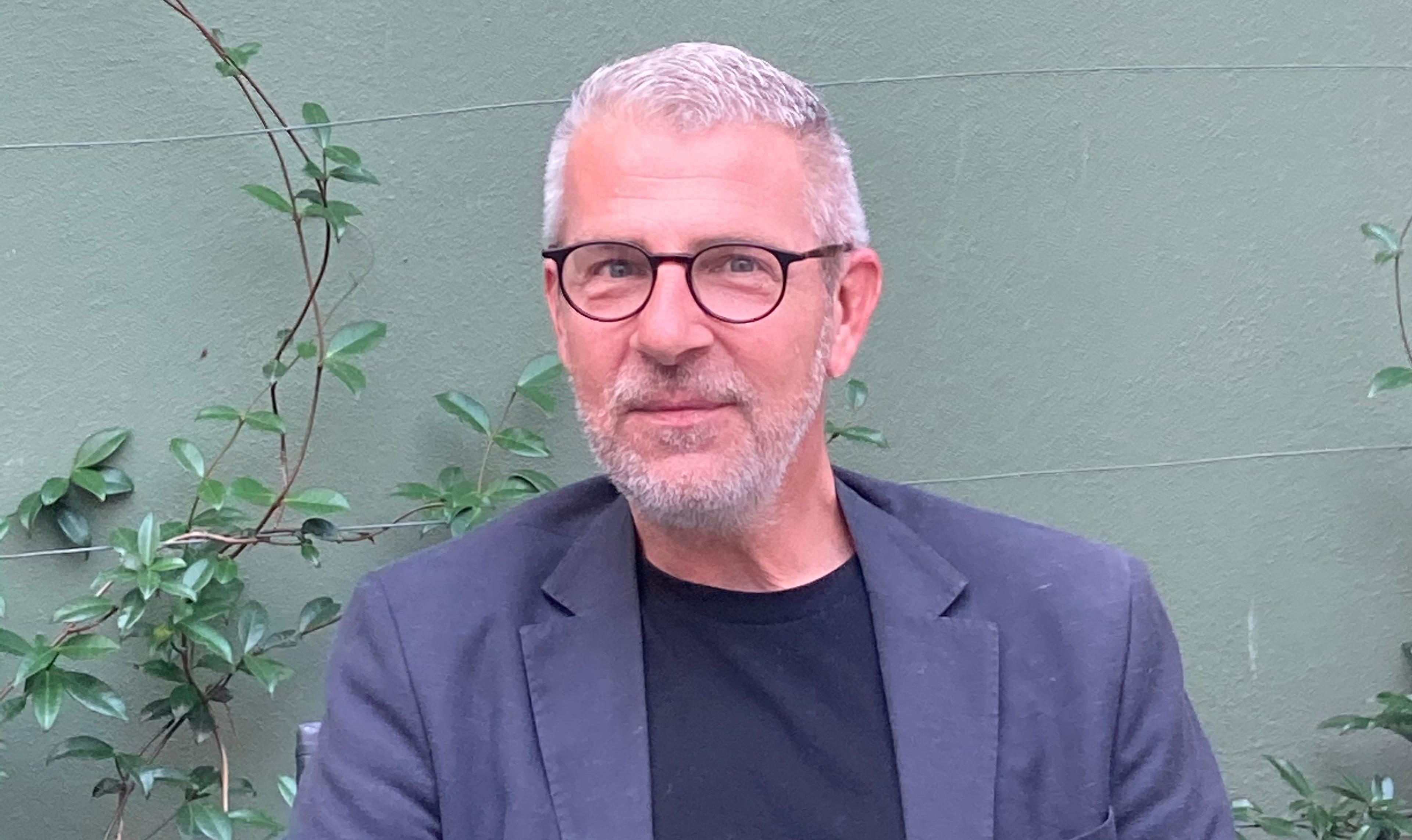 A man with grey hair and beard, wearing glasses, a black T-shirt, and a navy blazer, sits in front of a green wall adorned with climbing plants.