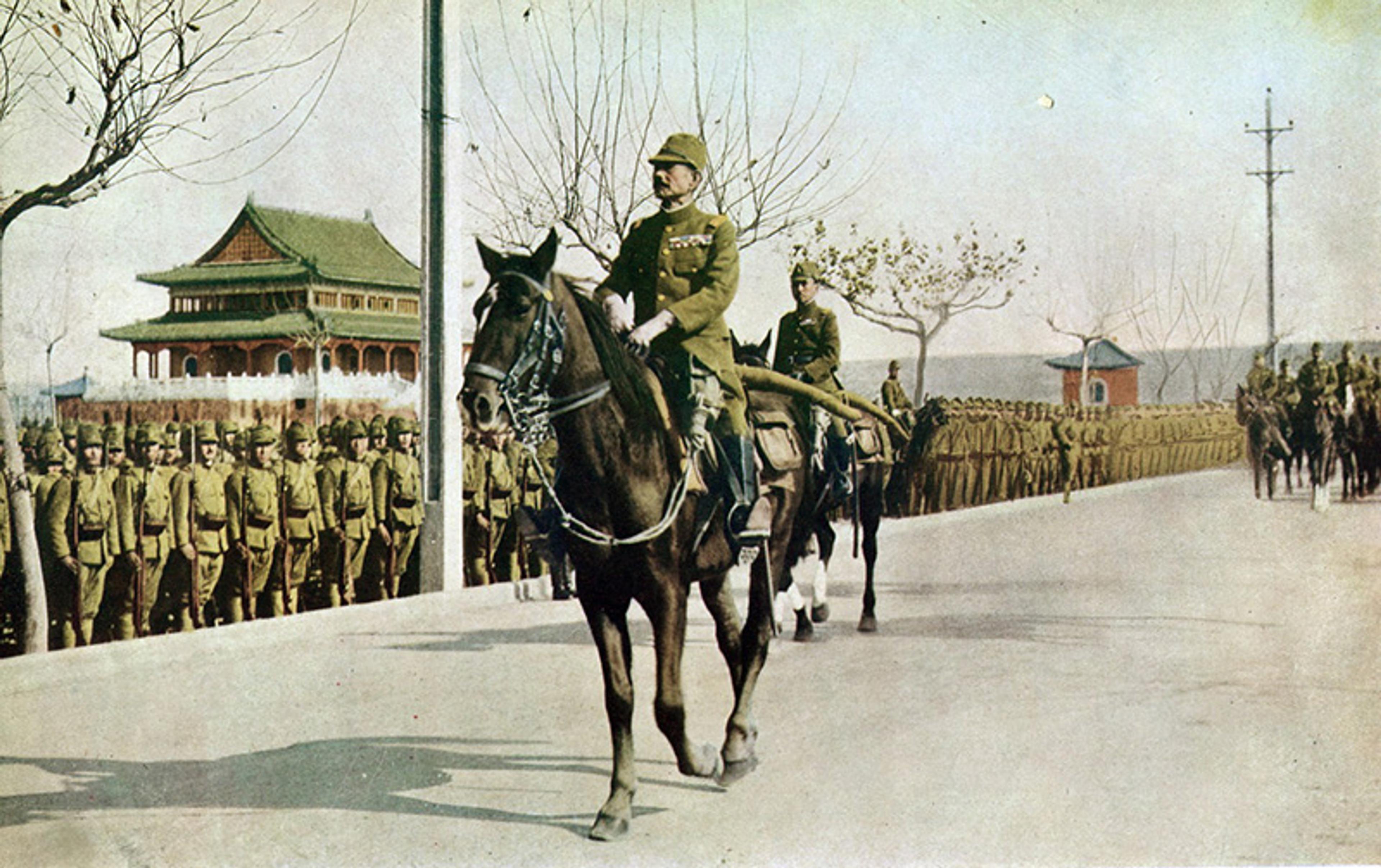 Soldiers in uniform march in formation with trees and telephone poles visible. In the foreground, a soldier on horseback leads the procession. A traditional building with a green, ornate roof is in the background, suggesting an Asian setting. The scene appears to be historical, possibly early 20th century.