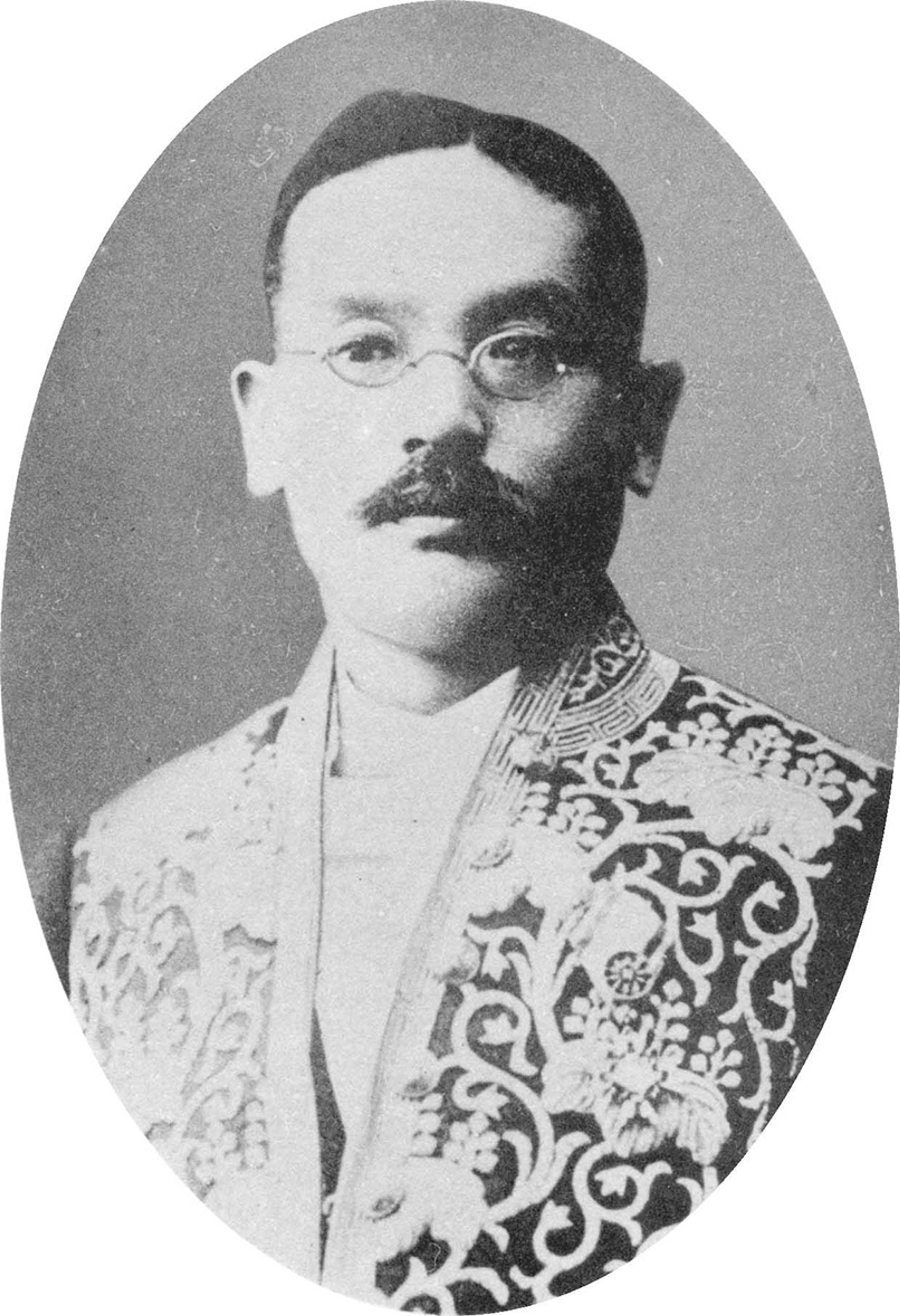 Black and white portrait of a man with a moustache and glasses, wearing an ornate, embroidered jacket with intricate floral patterns and animal motifs. The image is oval-shaped, with the man looking directly at the camera against a plain background.