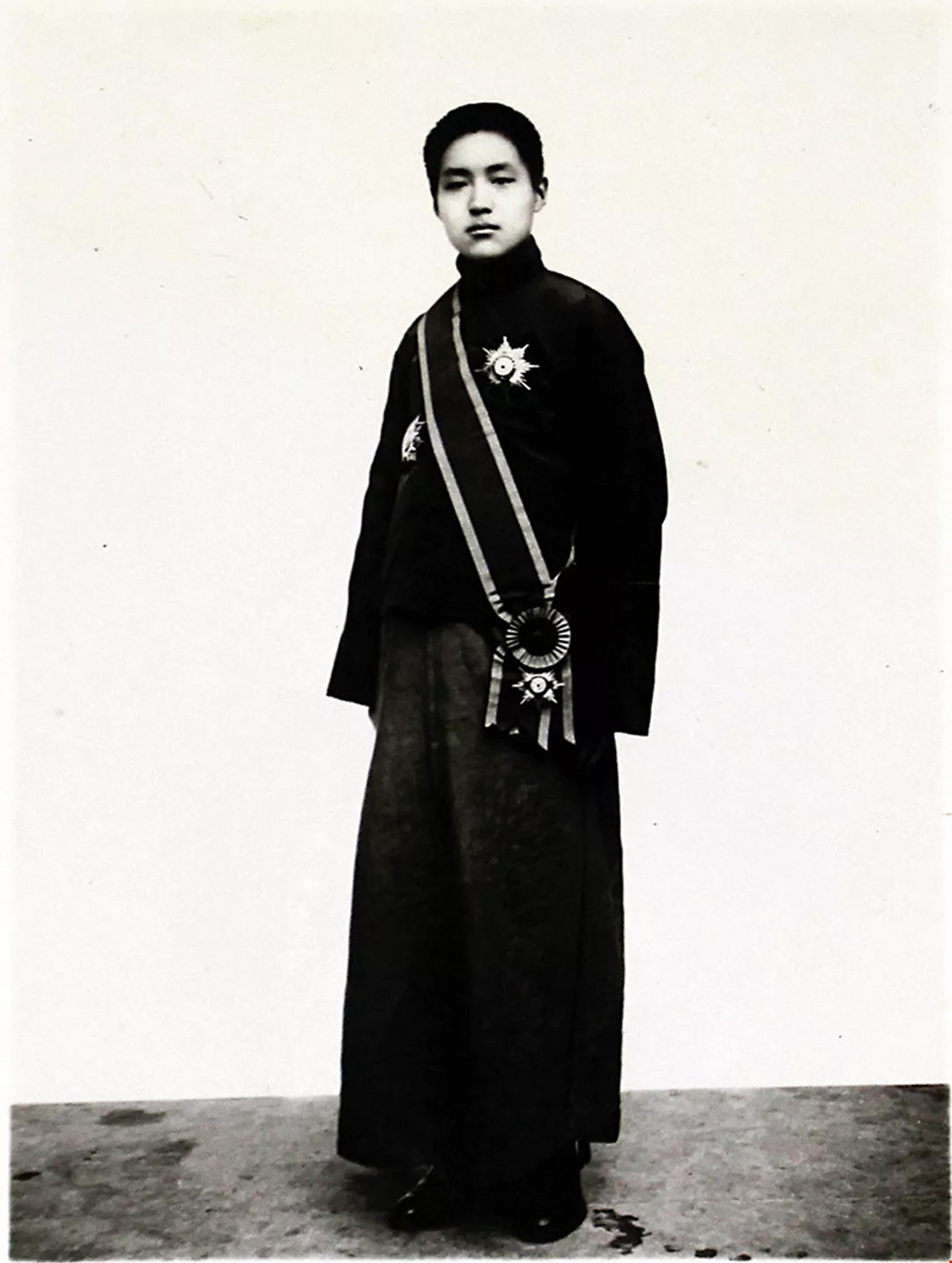 A young person standing in traditional attire, featuring a long robe and decorations including a sash and medals. The background is plain and light in colour, focusing attention entirely on the subject in the centre of the image.
