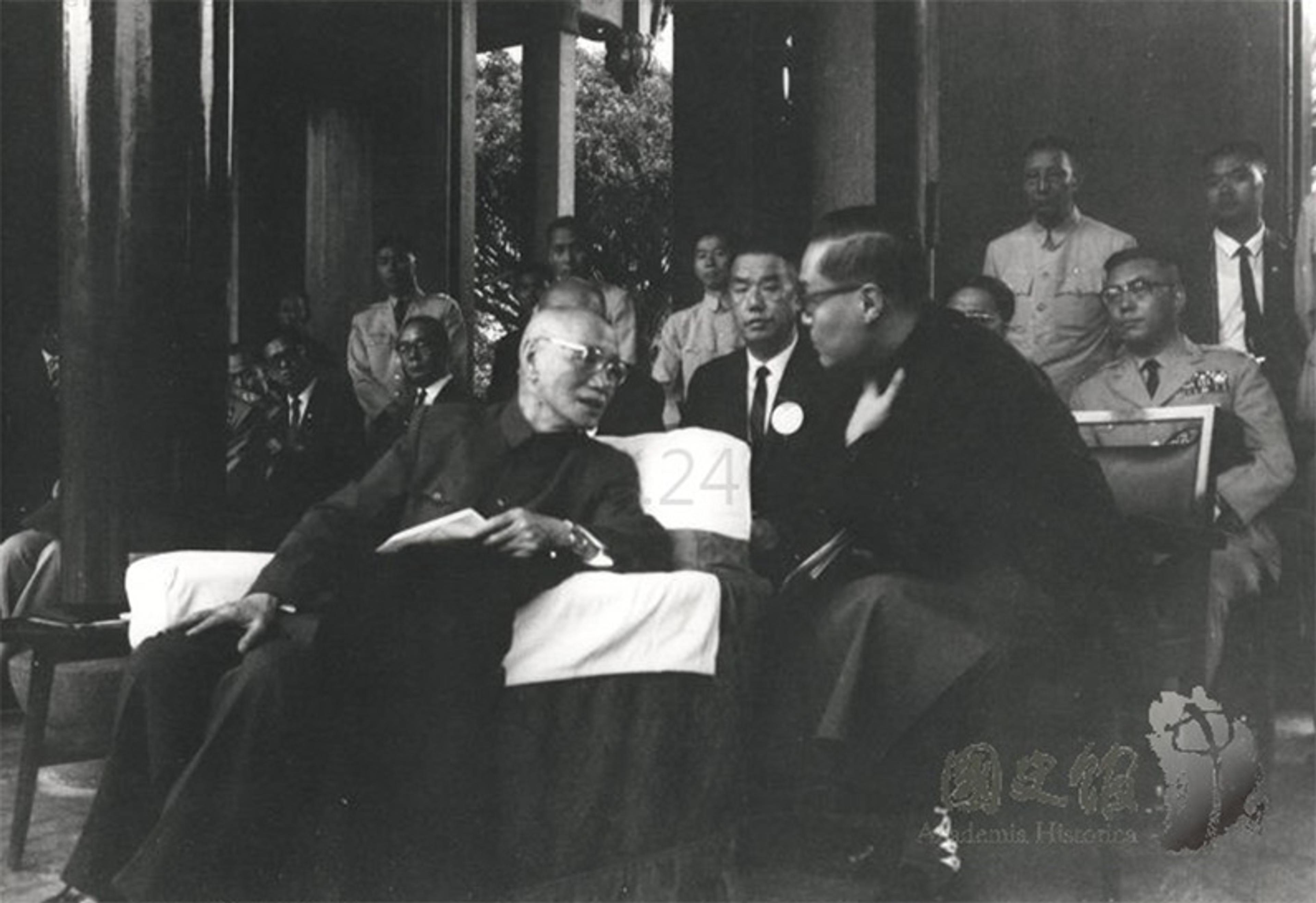 A black and white photo showing a group of men in a formal setting. In the foreground, one seated man is leaning forward, engaged in conversation with another. Other attendees in suits or uniforms can be seen standing in the background.