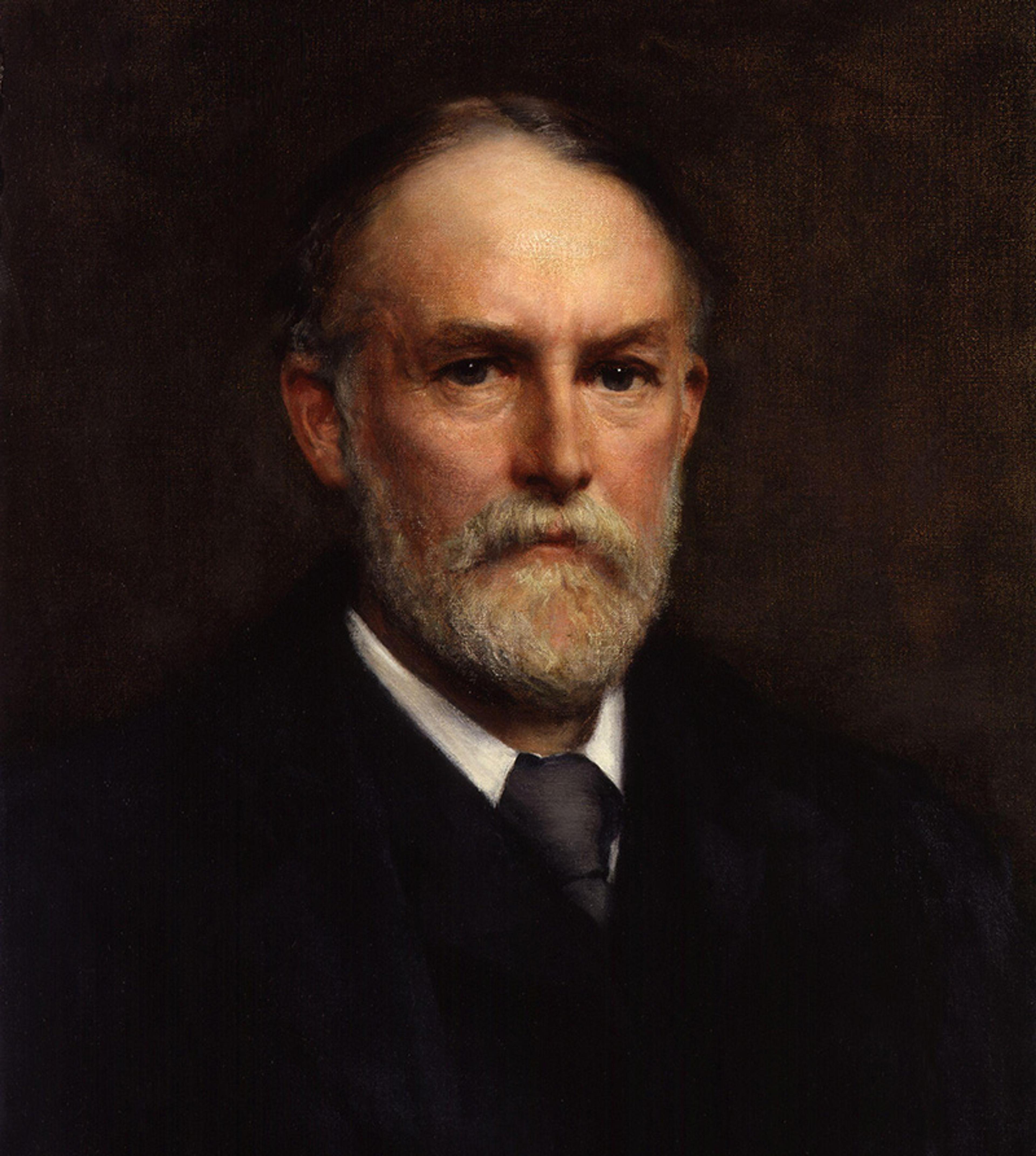Portrait of an older man with a beard, wearing a dark suit and tie, against a dark background.