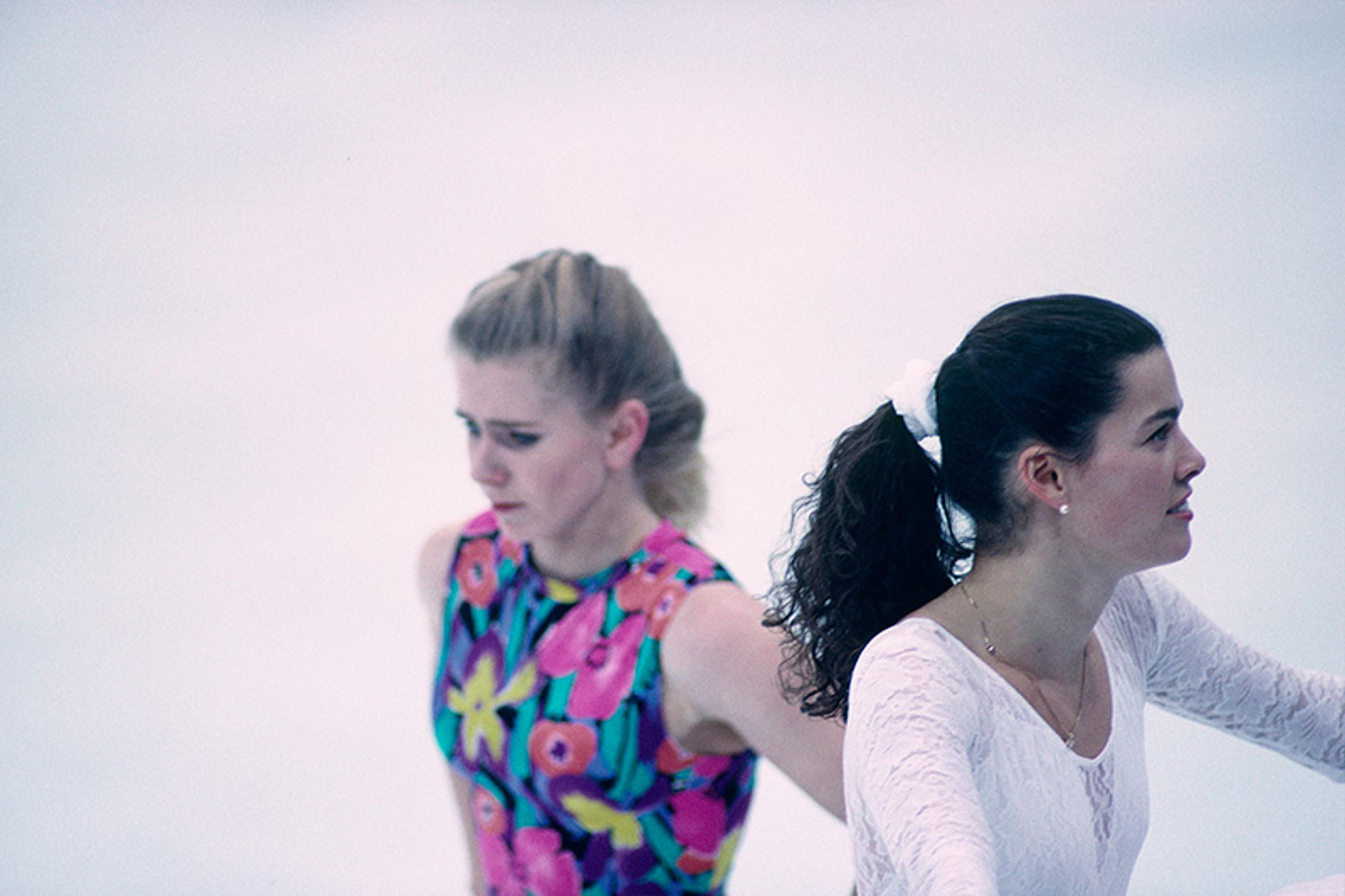 Two female ice skaters performing; one in a floral costume and the other in a white outfit, both concentrating on their routines on the ice rink.
