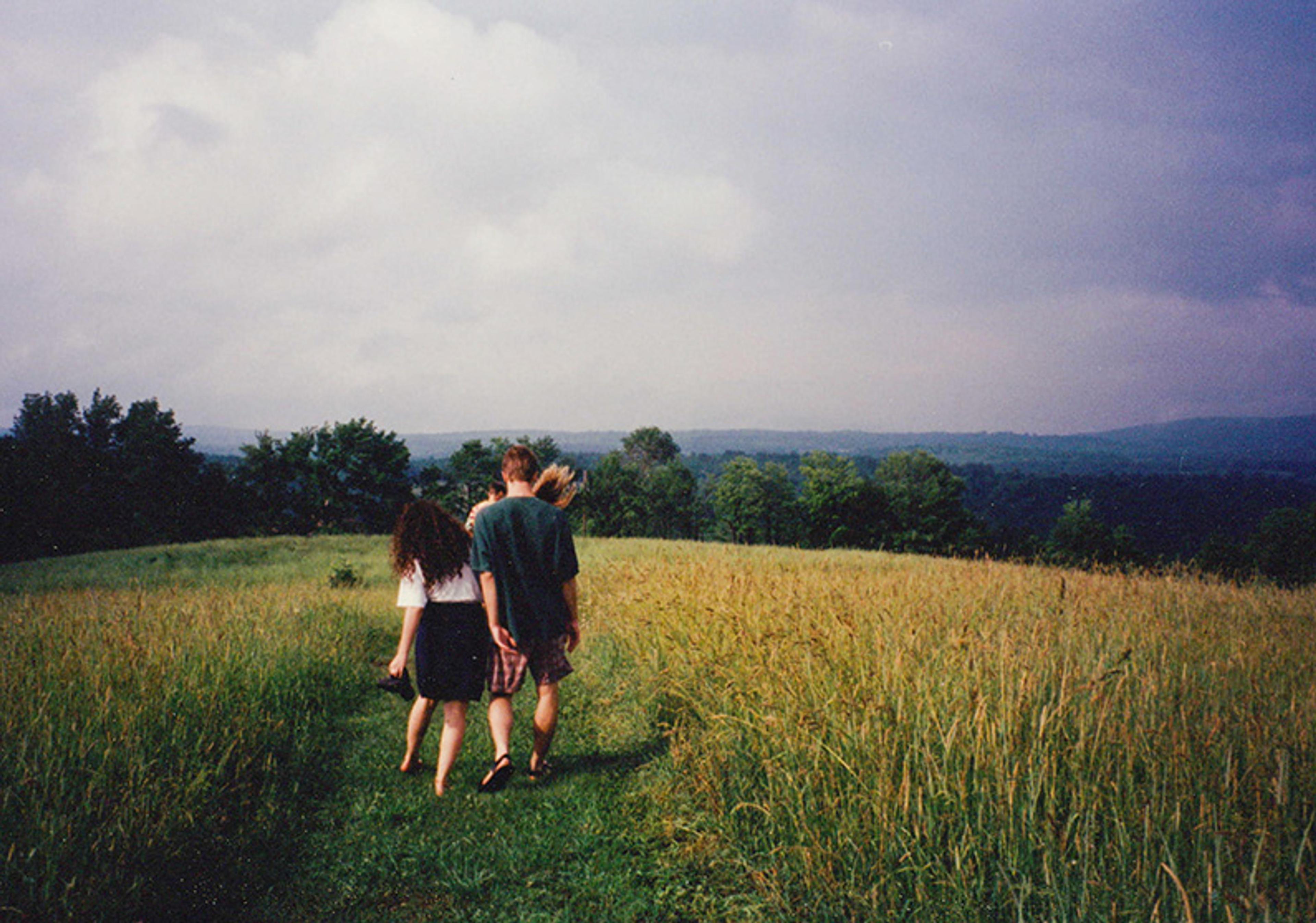 Three people walking on a path through a grassy field towards a forest under a cloudy sky.
