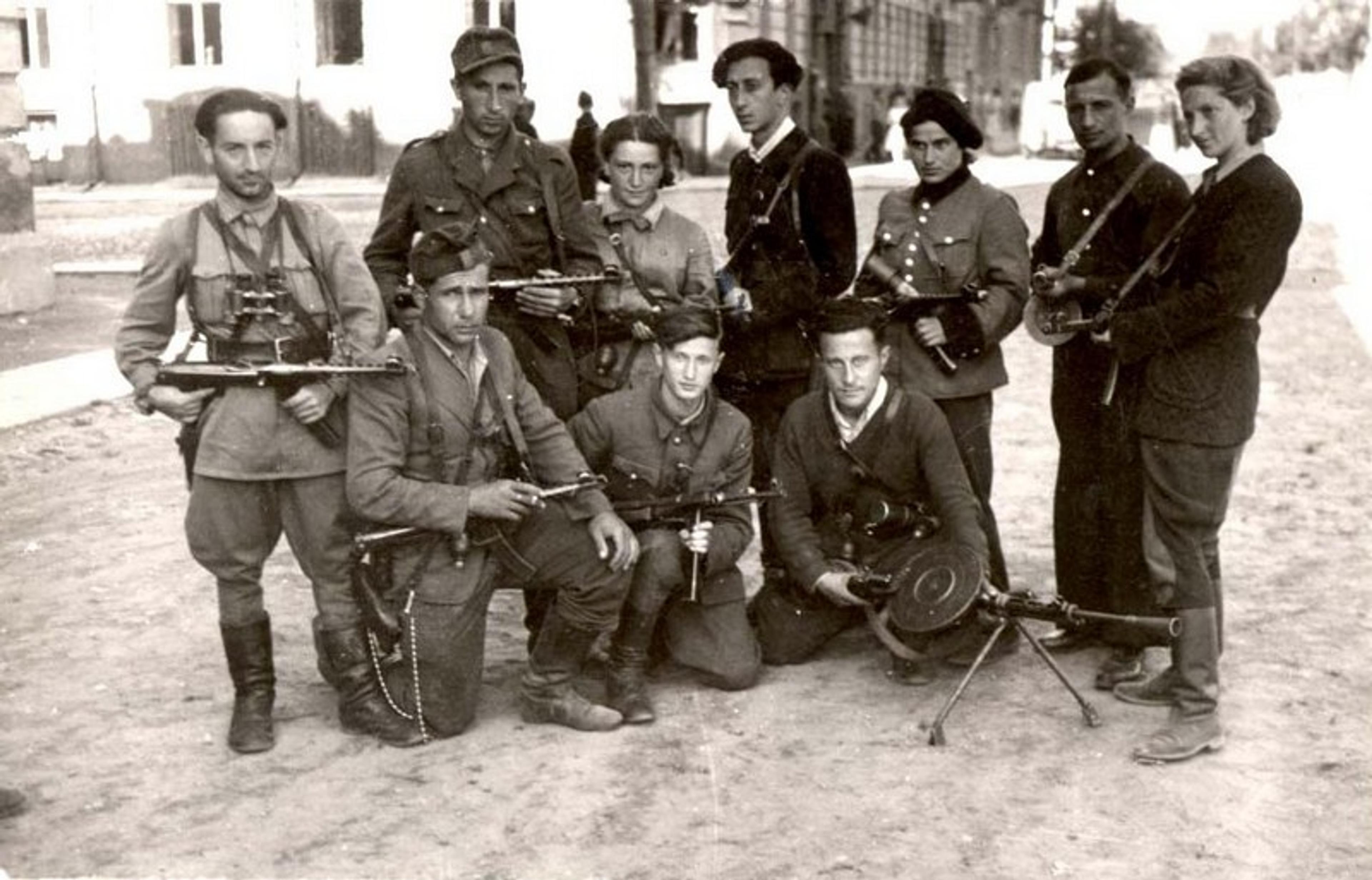 Group of nine armed individuals in military attire posing outdoors on a street during daytime, some crouching, others standing with weapons.