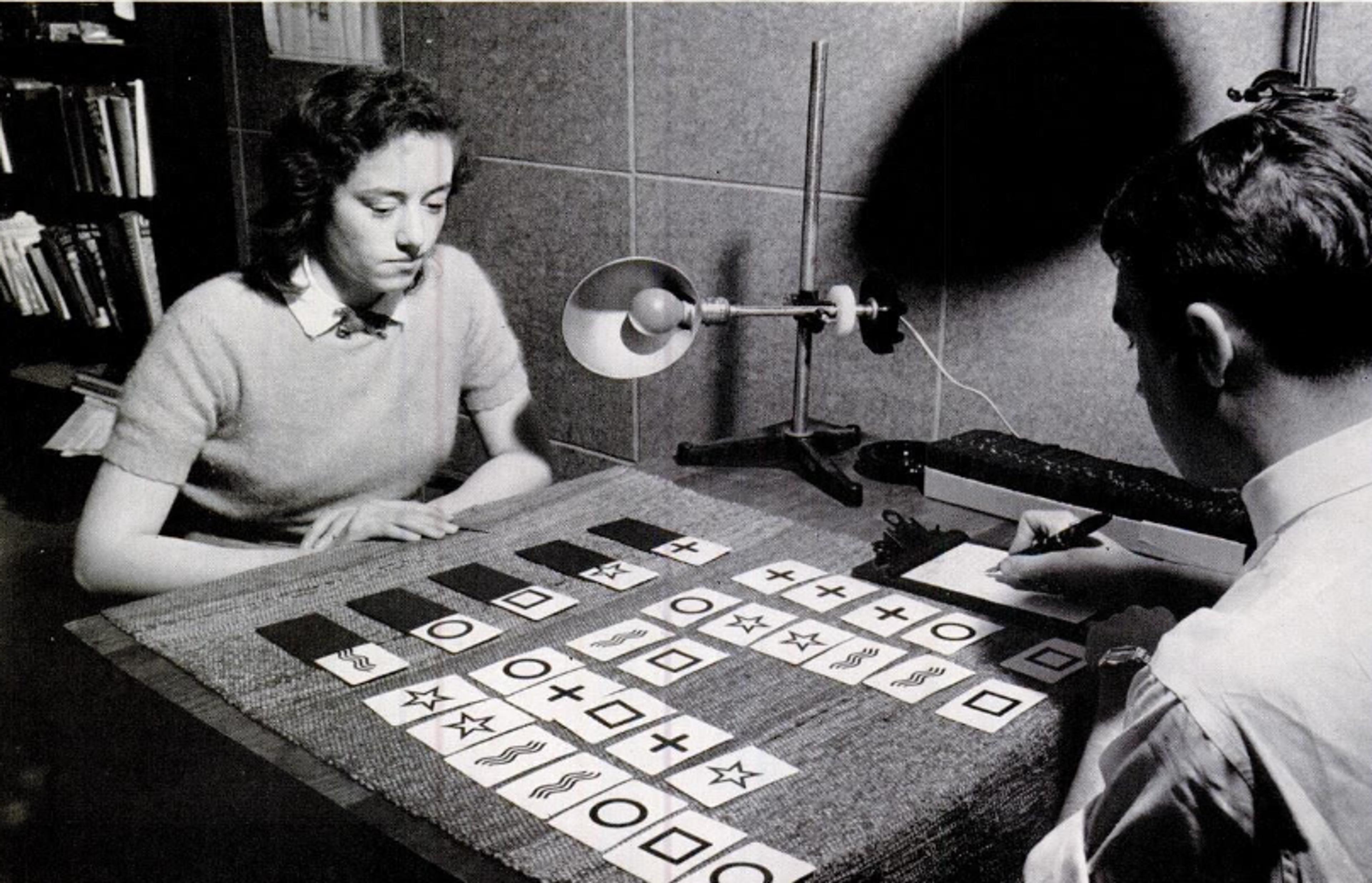 A woman and a man sit across from each other at a table, with cards displaying shapes and symbols laid out between them, engaging in a possible experiment.