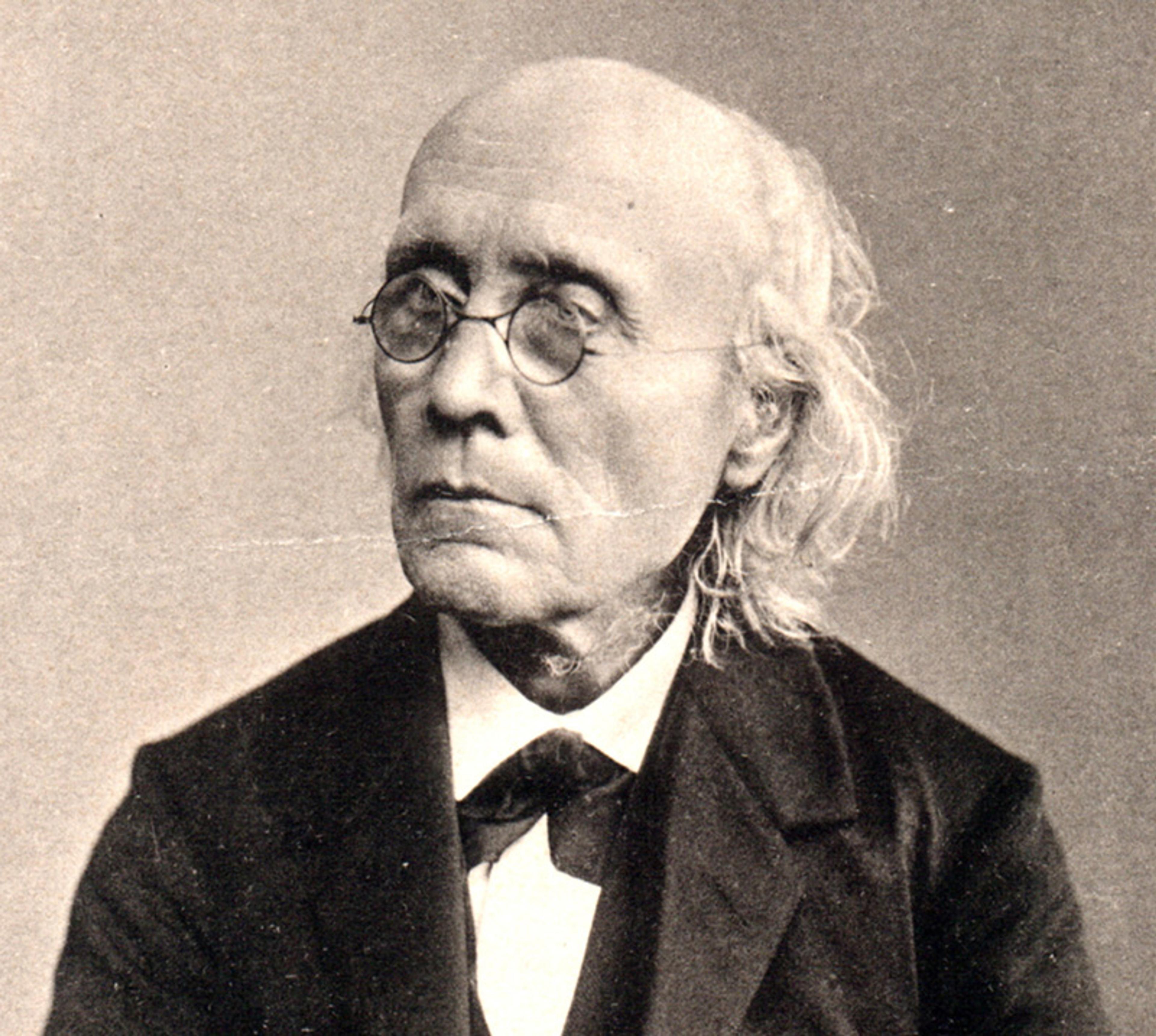 Sepia-toned portrait of an elderly man with glasses, a bald head, long hair, and dressed in formal clothing.