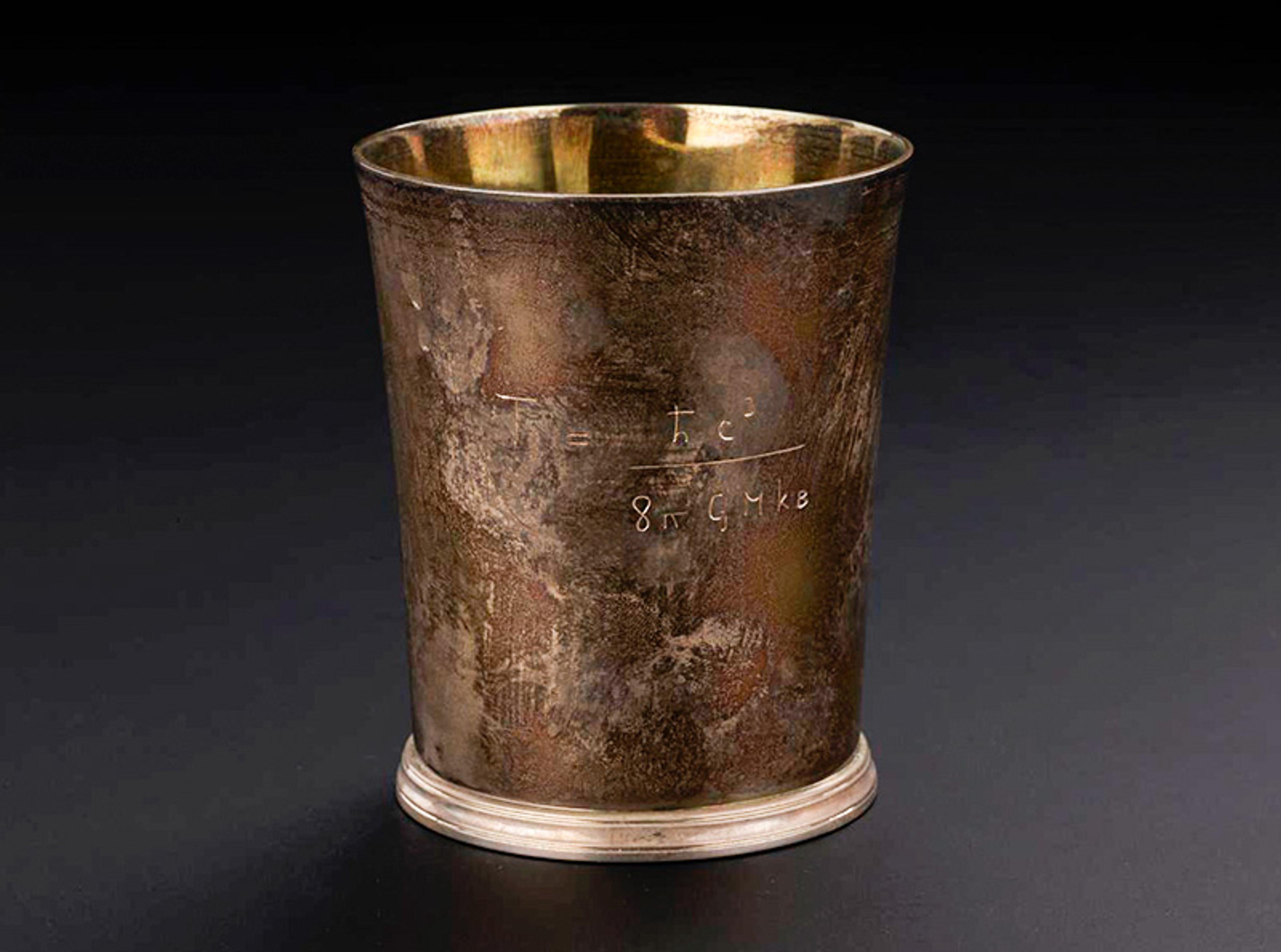 A metallic cylindrical cup with a tarnished, rustic appearance. The cup has a mathematical equation etched into its side. The background is black, highlighting the cup’s worn texture and showing its reflective golden interior.