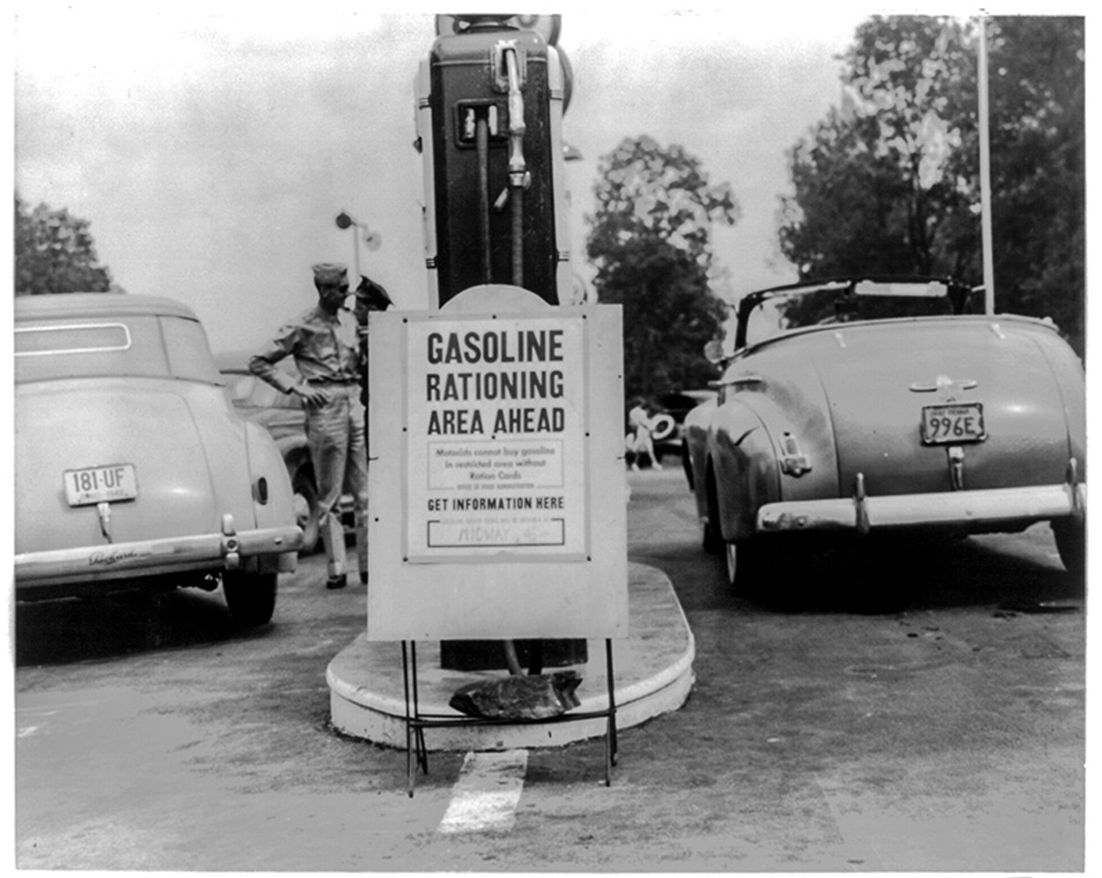 A black-and-white image showing a sign reading “Gasoline Rationing Area Ahead” with a petrol pump in the background. Two vintage cars are seen parked on either side of the sign, with a person in military uniform standing near the pump. Trees are visible in the background.