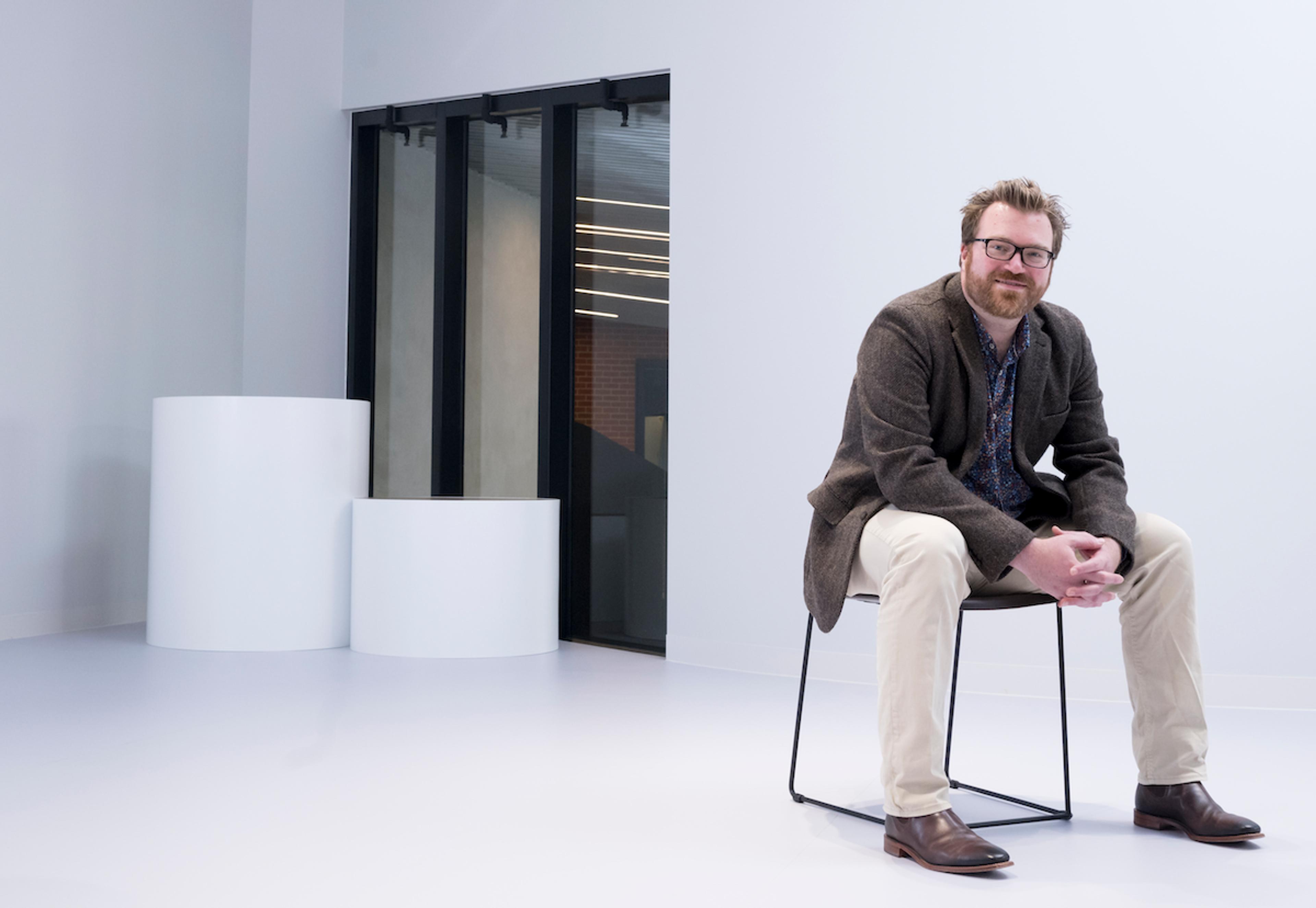 A man with glasses, wearing a brown blazer, sits on a chair in a minimalist white room with large windows and cylindrical sculptures.