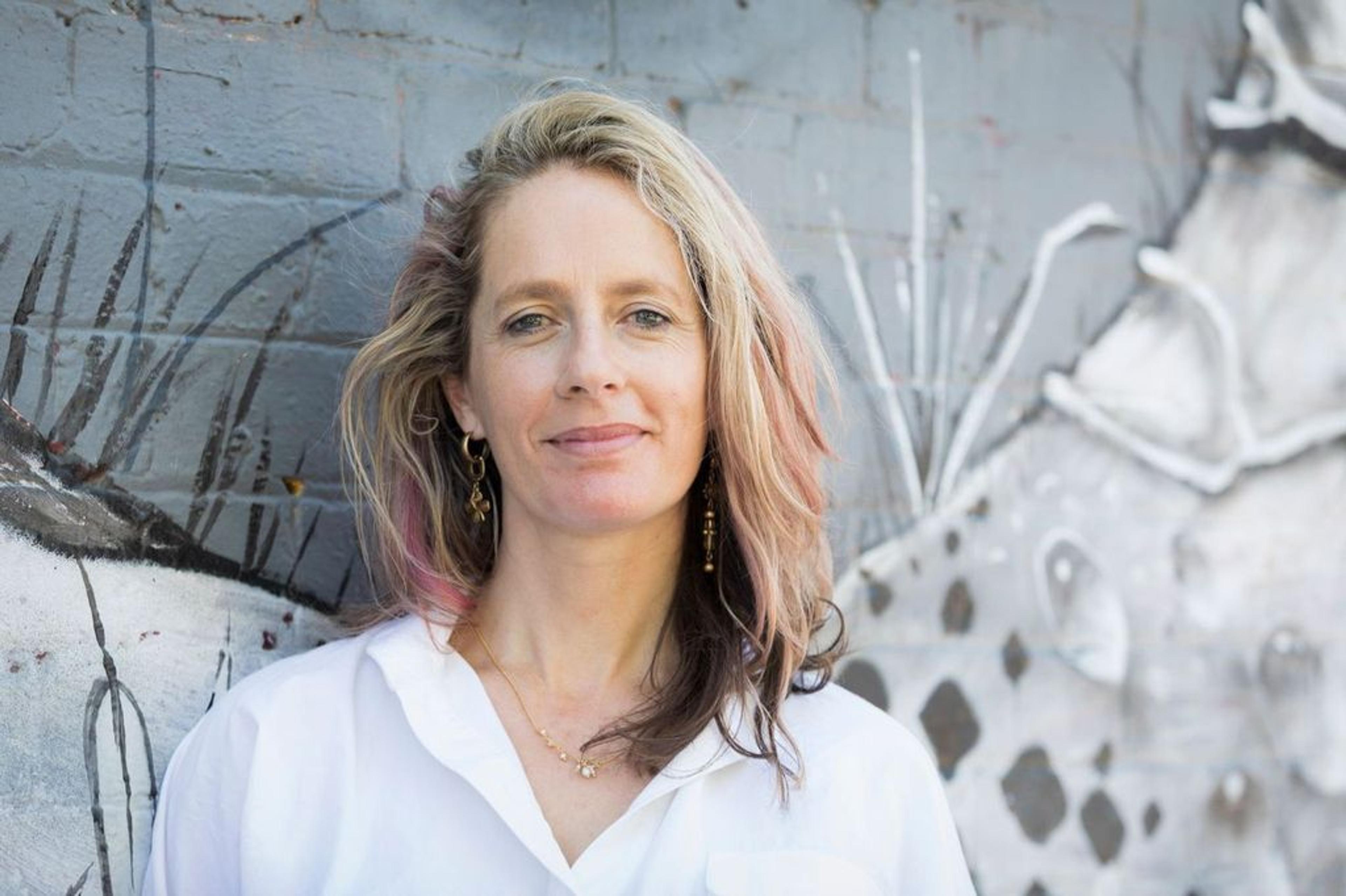 A woman with blonde hair and highlights, wearing a white shirt, stands in front of a mural on a brick wall.