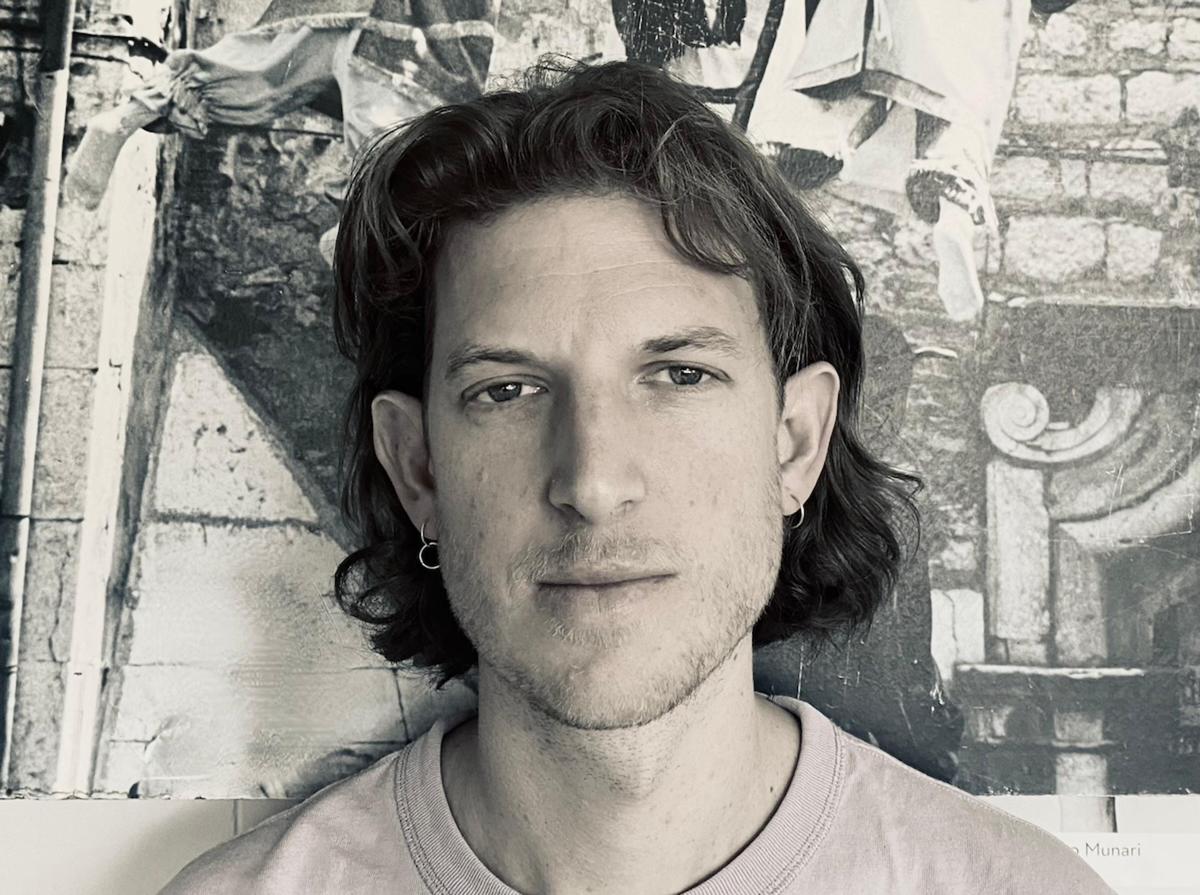 A person with shoulder-length hair, wearing earrings and a light-coloured shirt, stands in front of a black and white historical mural.