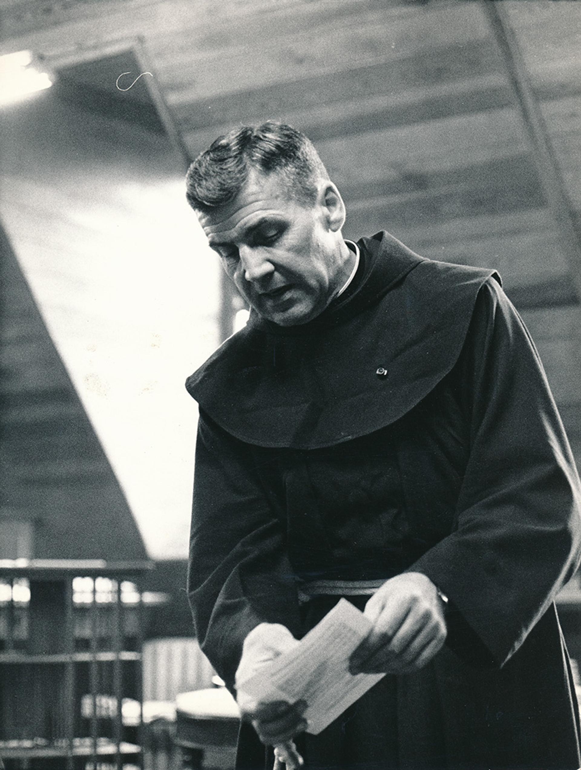 A black-and-white photo of a male priest in a hooded robe, holding a sheet of paper, standing in a room with wooden walls and shelves in the background.