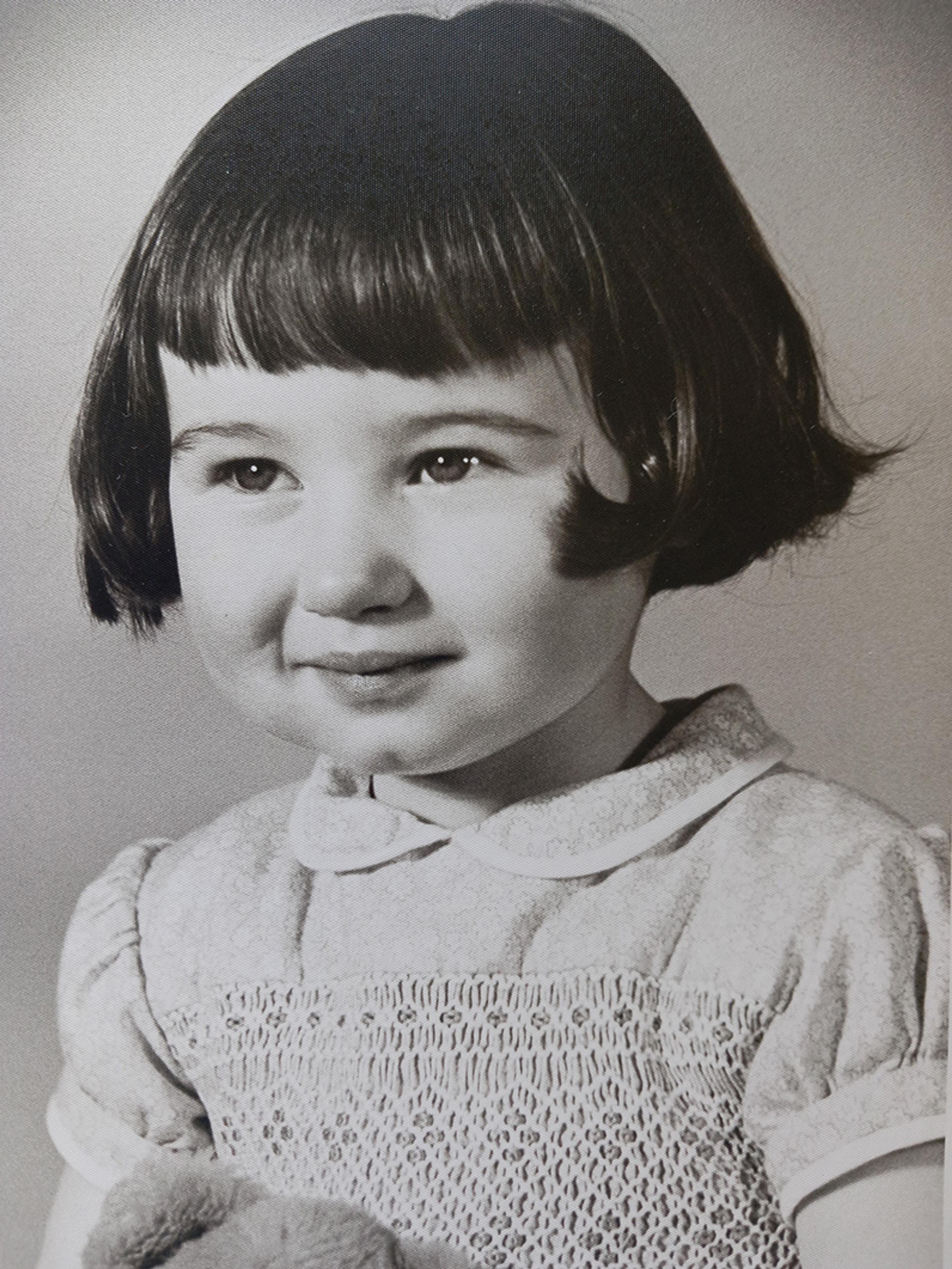 Black-and-white portrait of a young girl with short dark hair and a fringe, wearing a collared dress, holding a soft toy.