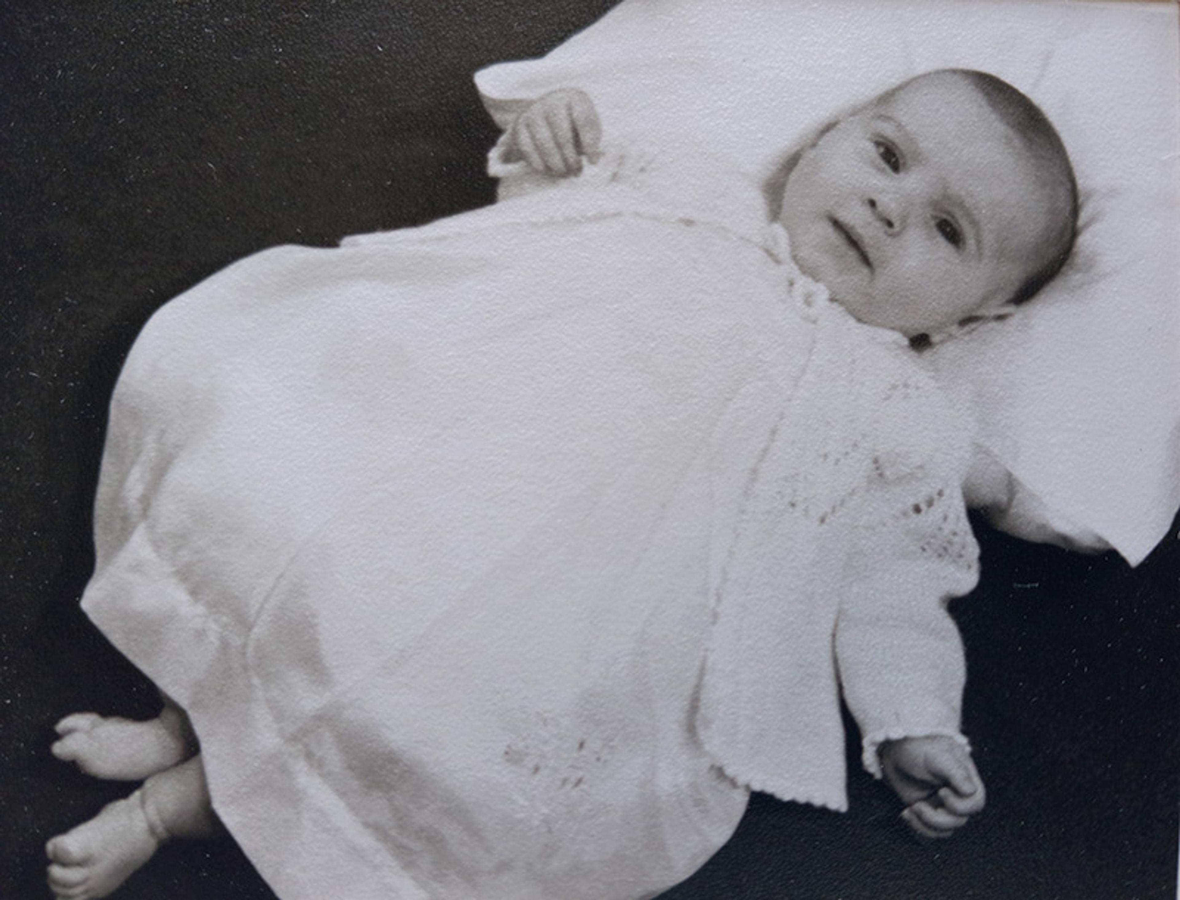Black-and-white photo of a baby lying on a cushion, wearing a white christening gown and smiling slightly.