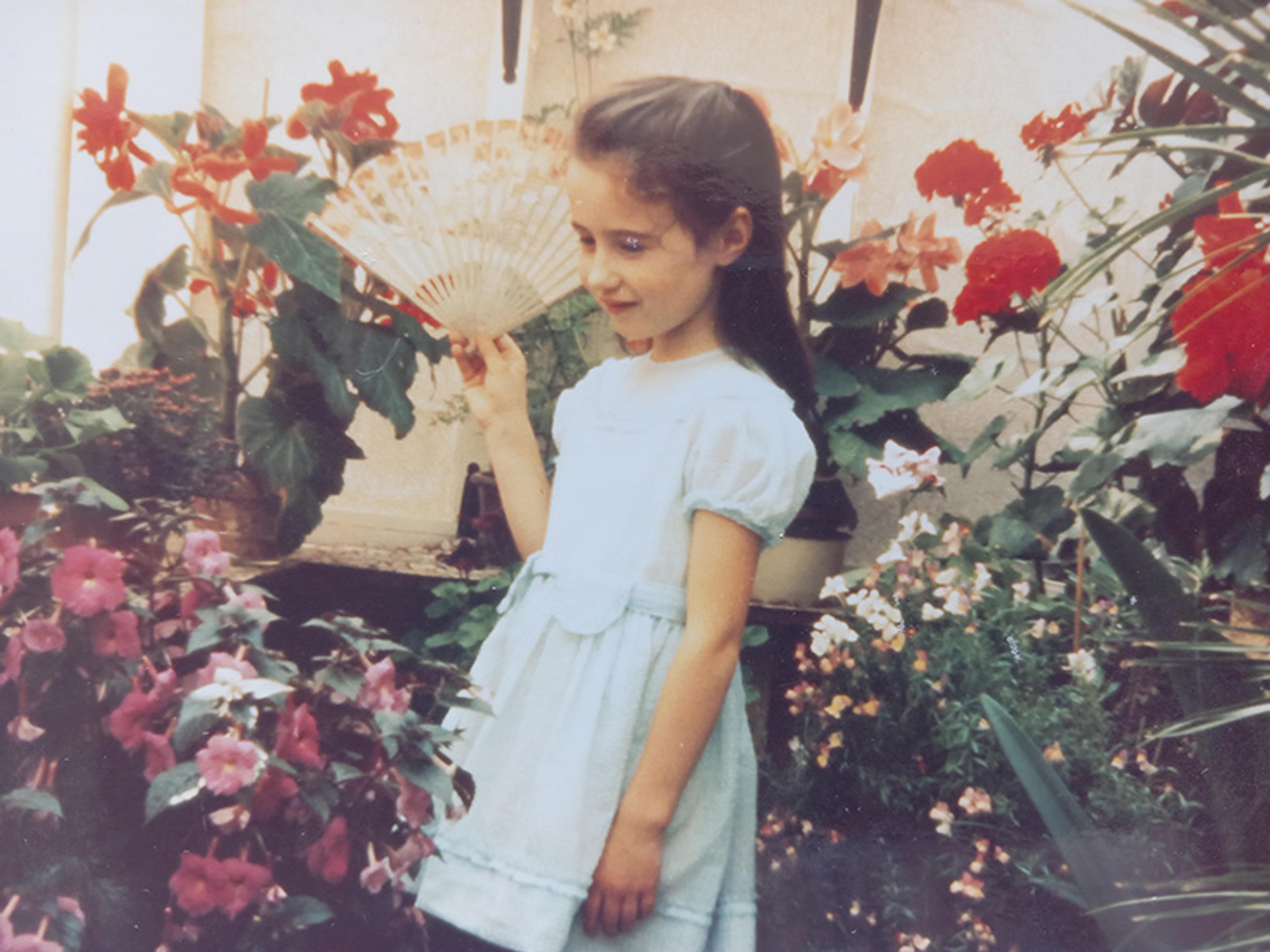 A young girl in a light blue dress holds a fan, standing among colourful flowers in a garden.