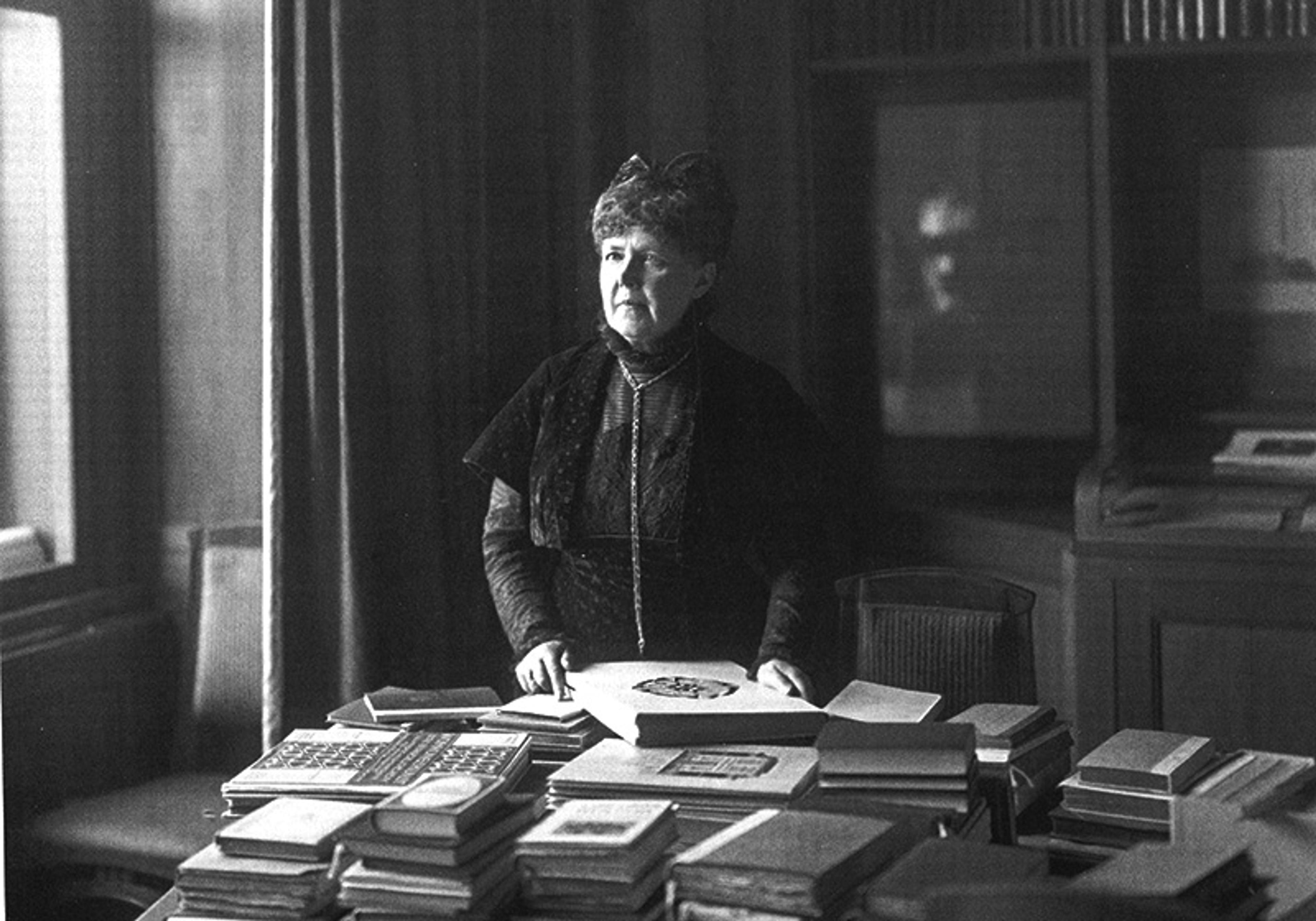 An elderly woman stands behind a desk filled with stacks of books in a dimly lit room, wearing a dark dress and looking towards a window.