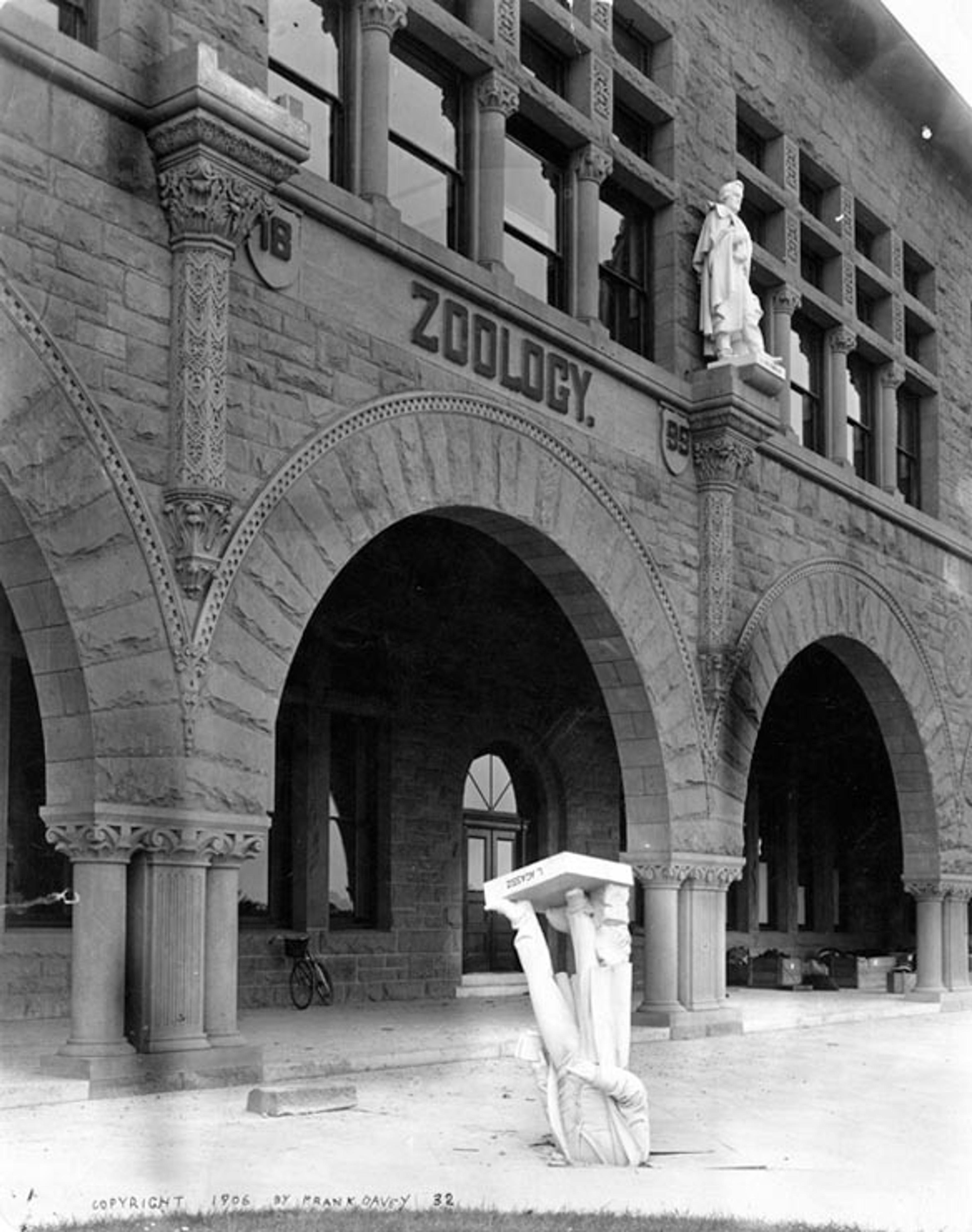 Archways of a Zoology building from 1899 shown behind a toppled statue of a figure standing on its head. The building has gothic architectural elements.