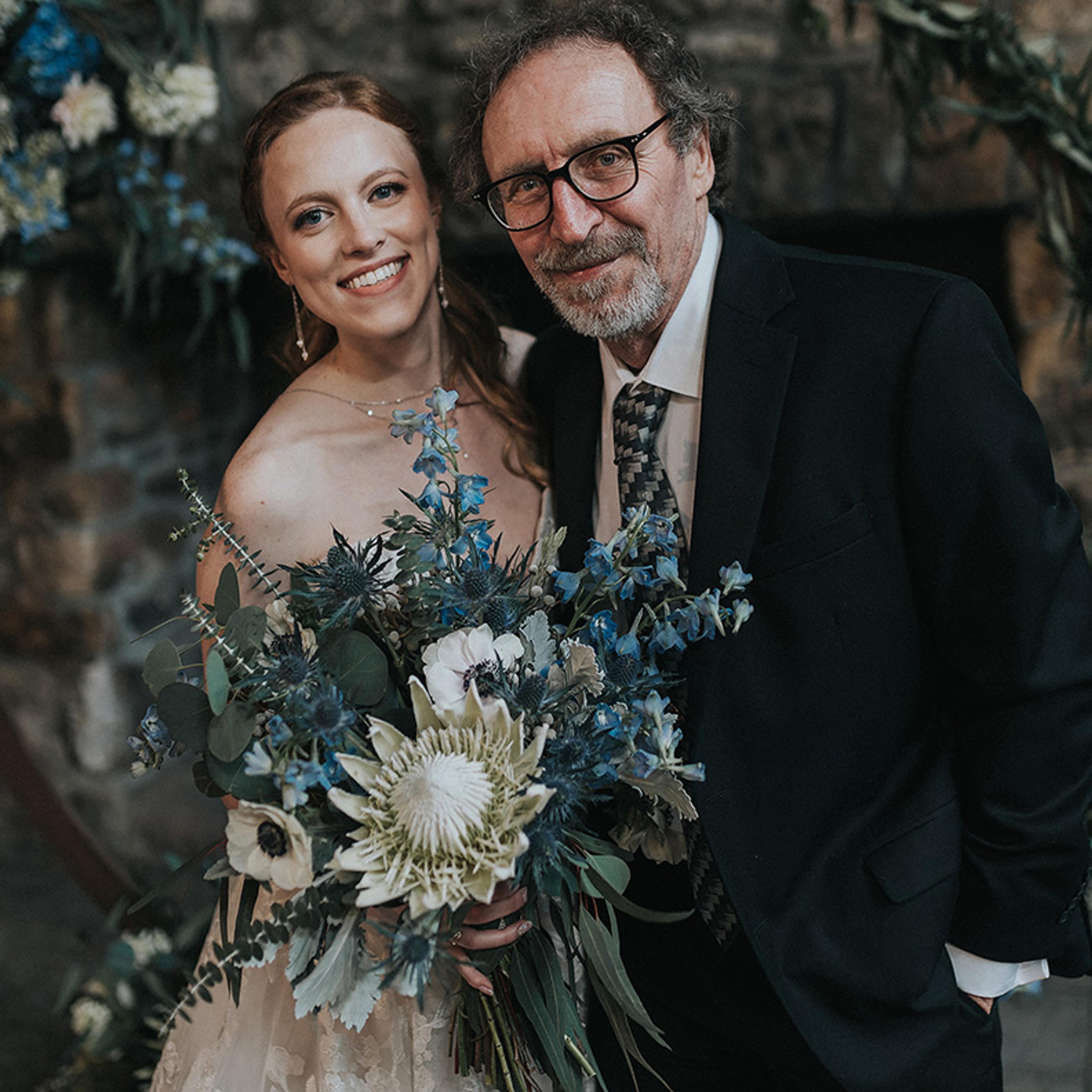 Bride in a wedding dress holds a bouquet of flowers and stands next to a man in a suit, both smiling.