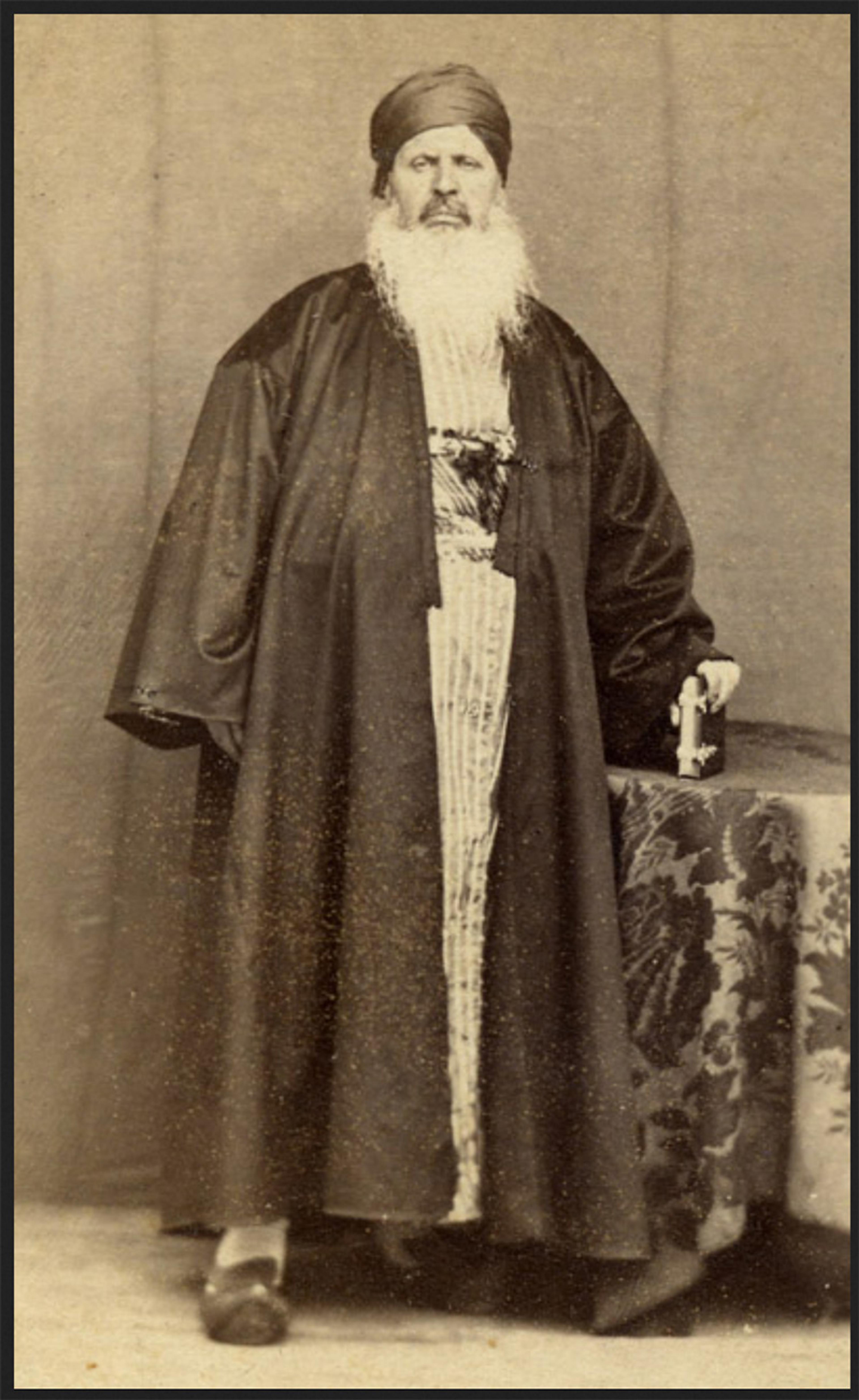 Sepia vintage photograph of a man with a long beard, wearing traditional robes and a head covering, standing beside a table with a patterned cloth.