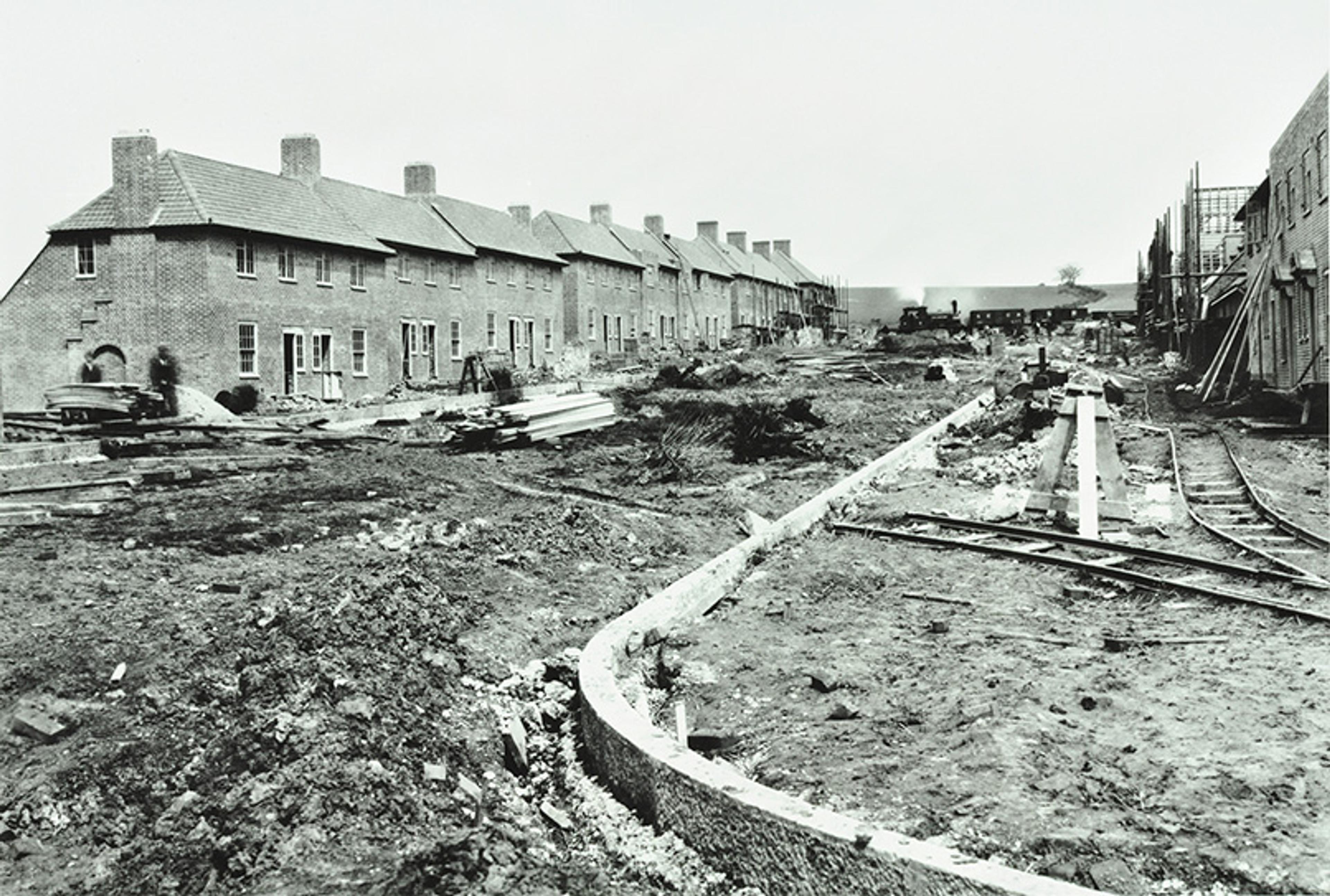 Black-and-white photo of a building site with a row of terraced houses on the left and building materials scattered around the dirt-covered ground.