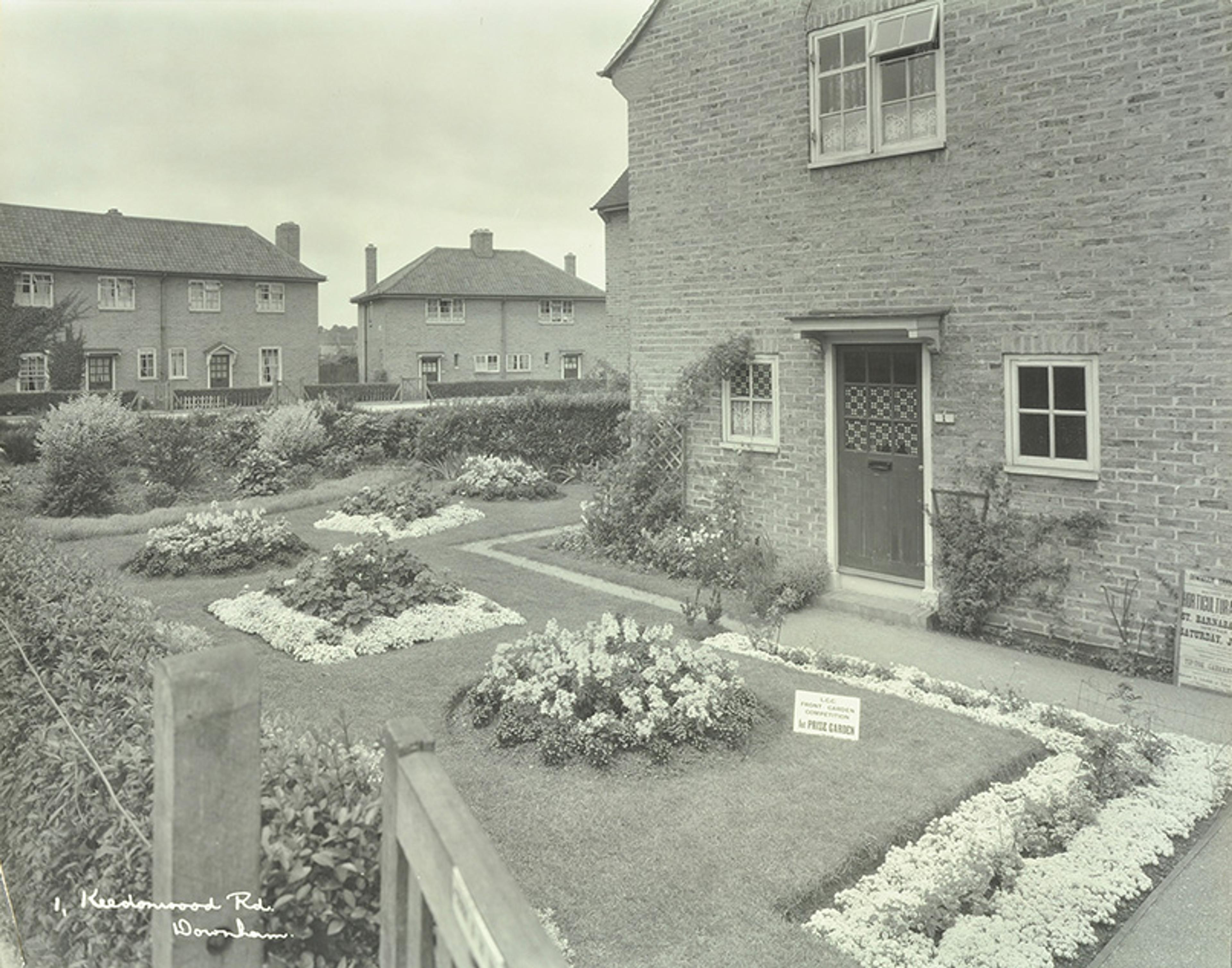 Black-and-white photo of front garden of a brick house with neat, rectangular flower beds and a sign reading “1st Prize Garden.” Two similar houses are visible in the background.