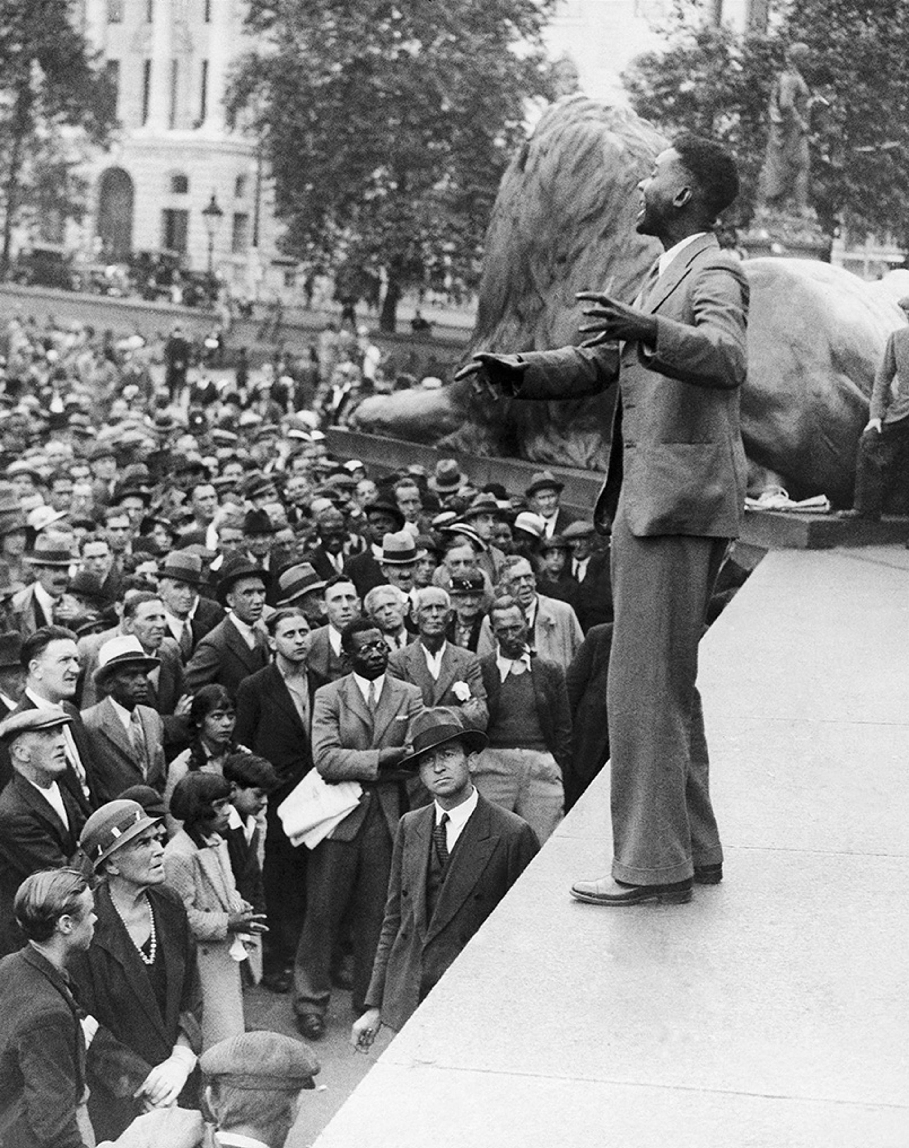 Black-and-white photo of a man speaking to a large crowd at Trafalgar Square with a lion statue visible in the background.