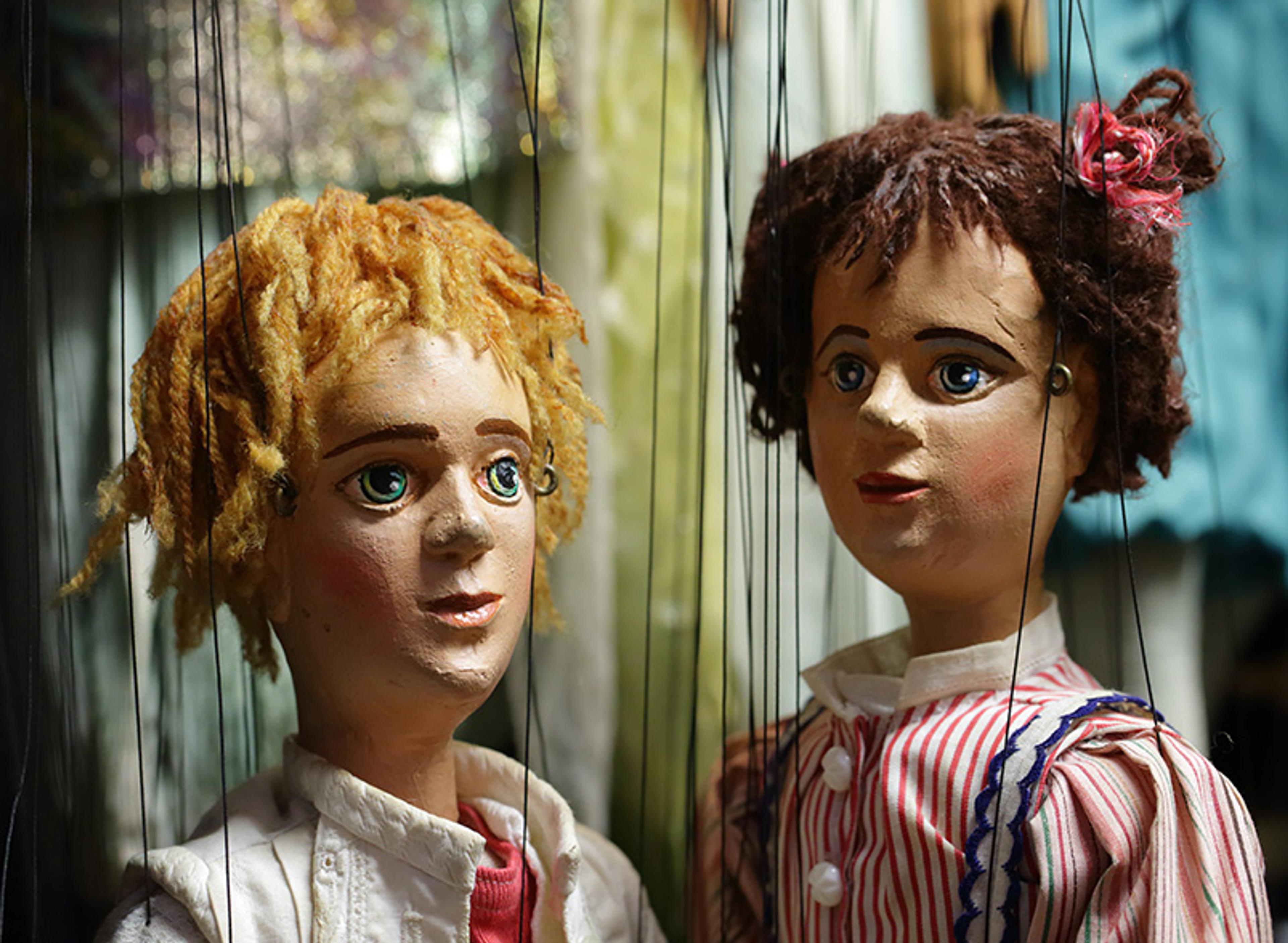 Photo of two marionette puppets with detailed facial features, one with blonde hair wearing white and the other with brown hair in striped clothing.