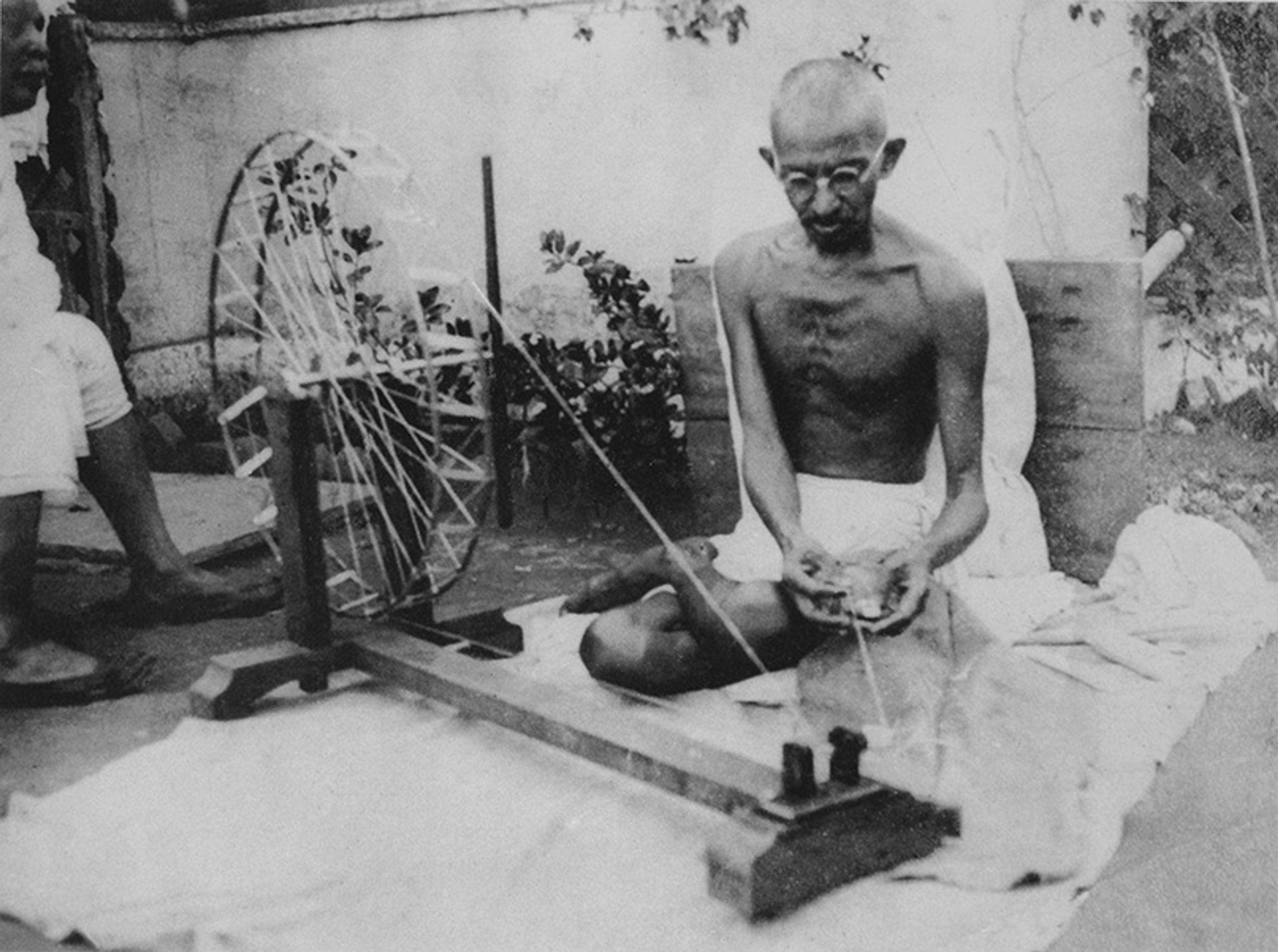 Black-and-white photo of a man sitting cross-legged on the ground using a spinning wheel, with another person partially visible.