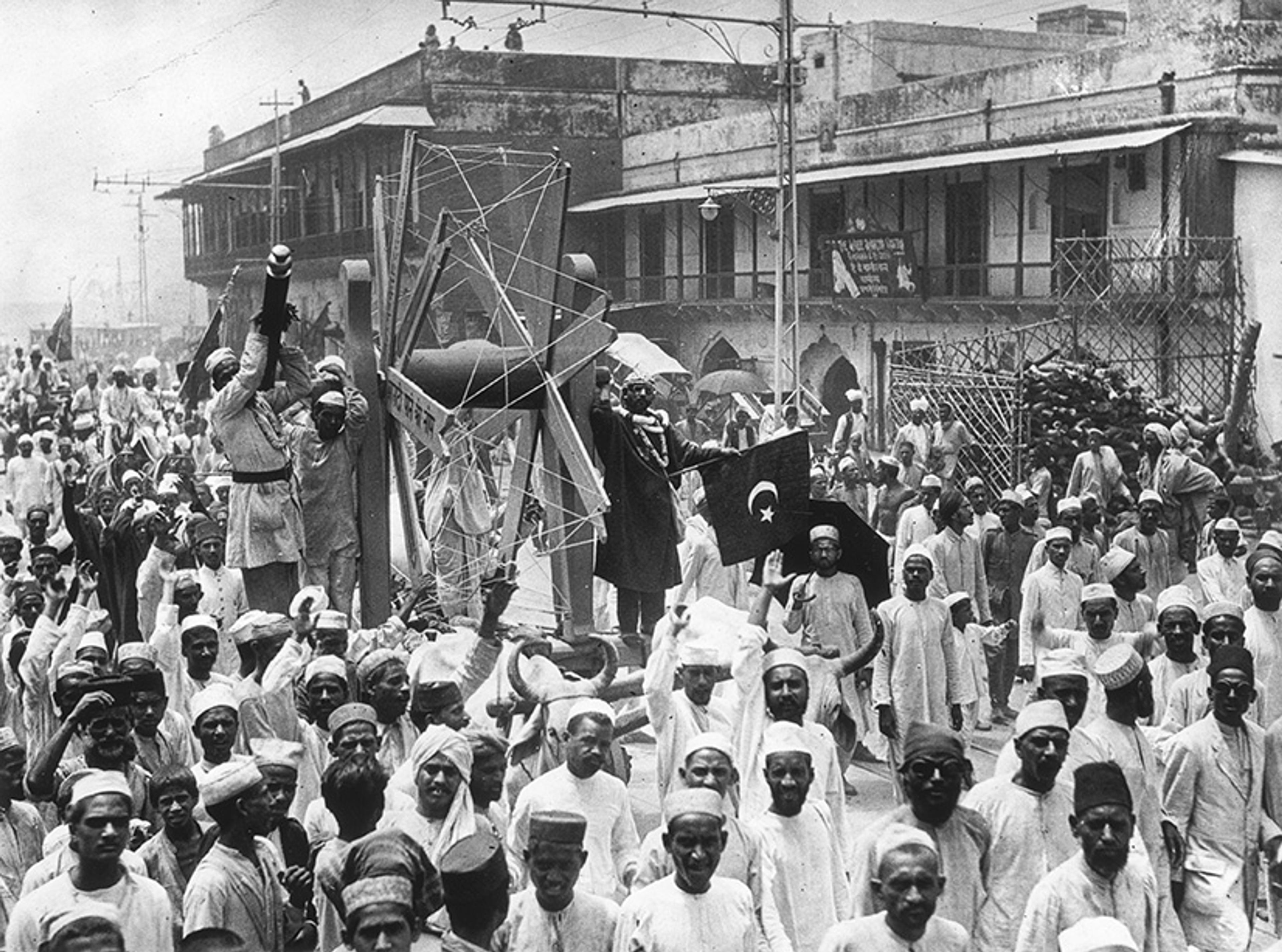 Black and white photo of a large crowd in Indian attire in a street parade, featuring a spoked wooden structure and flags.