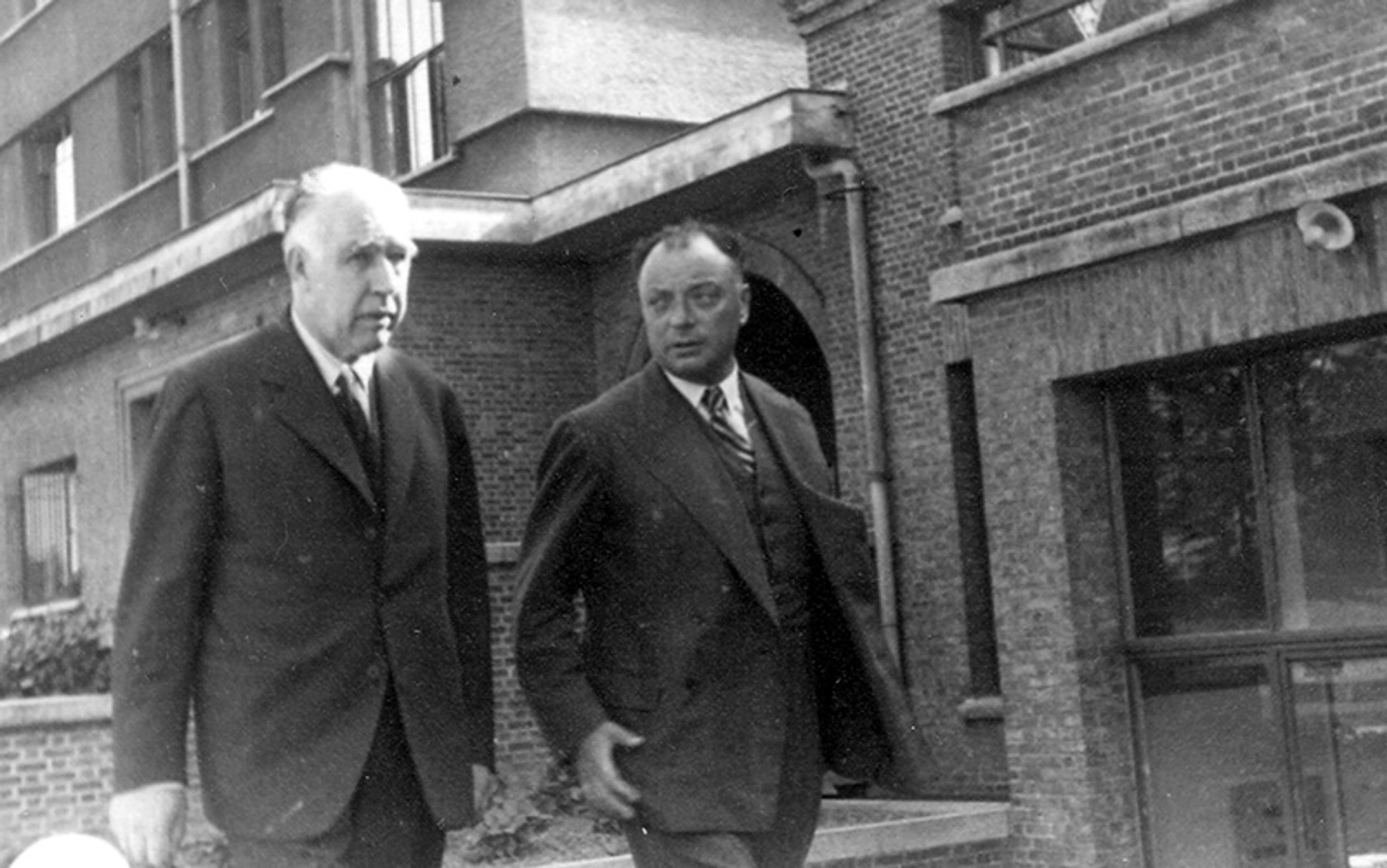 Black and white photo of two men in suits walking outside a brick building, engaging in conversation.