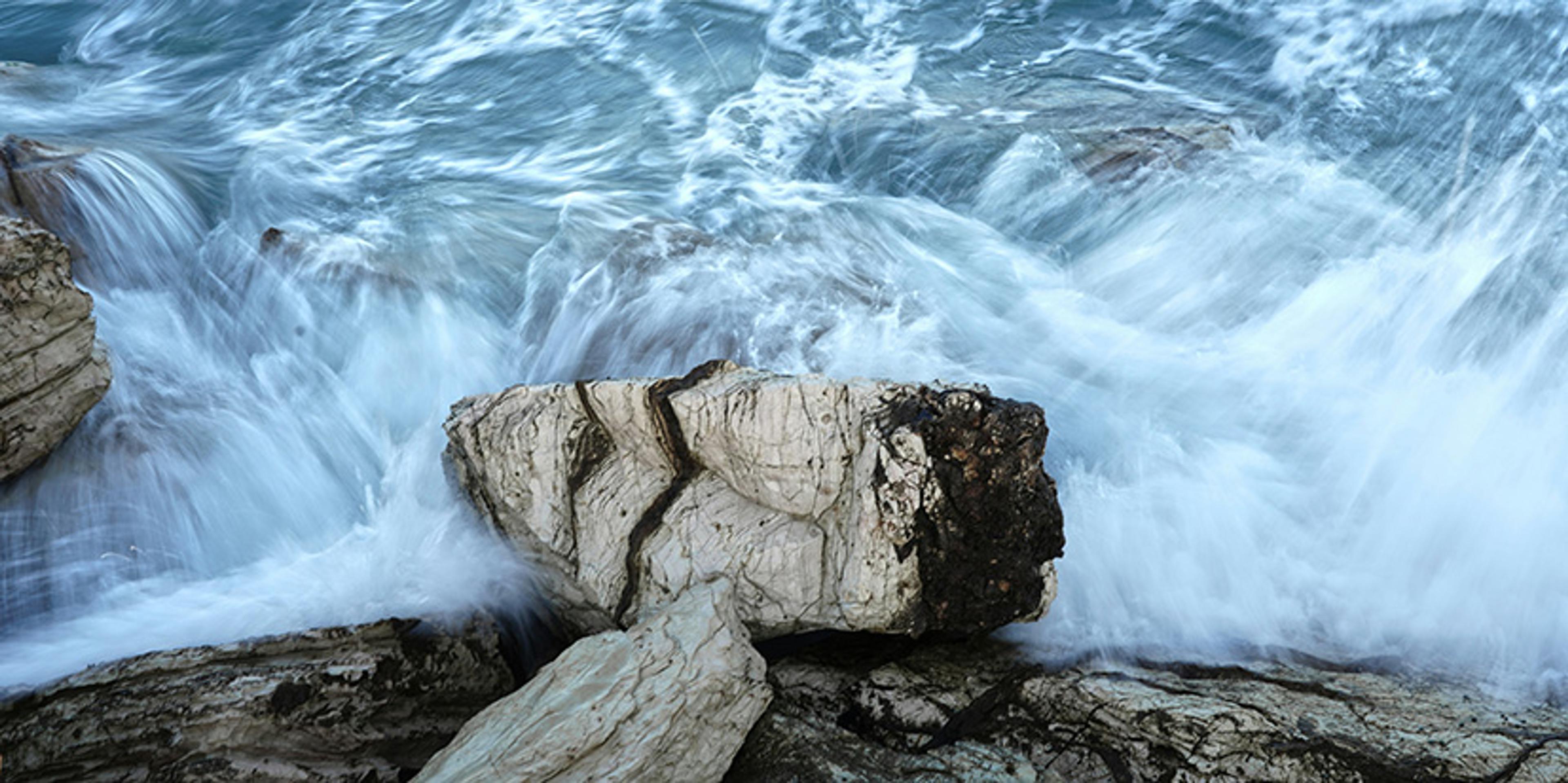 Photo of waves crashing over large rocks by the shoreline, capturing the movement of the water and the texture of the rocks.