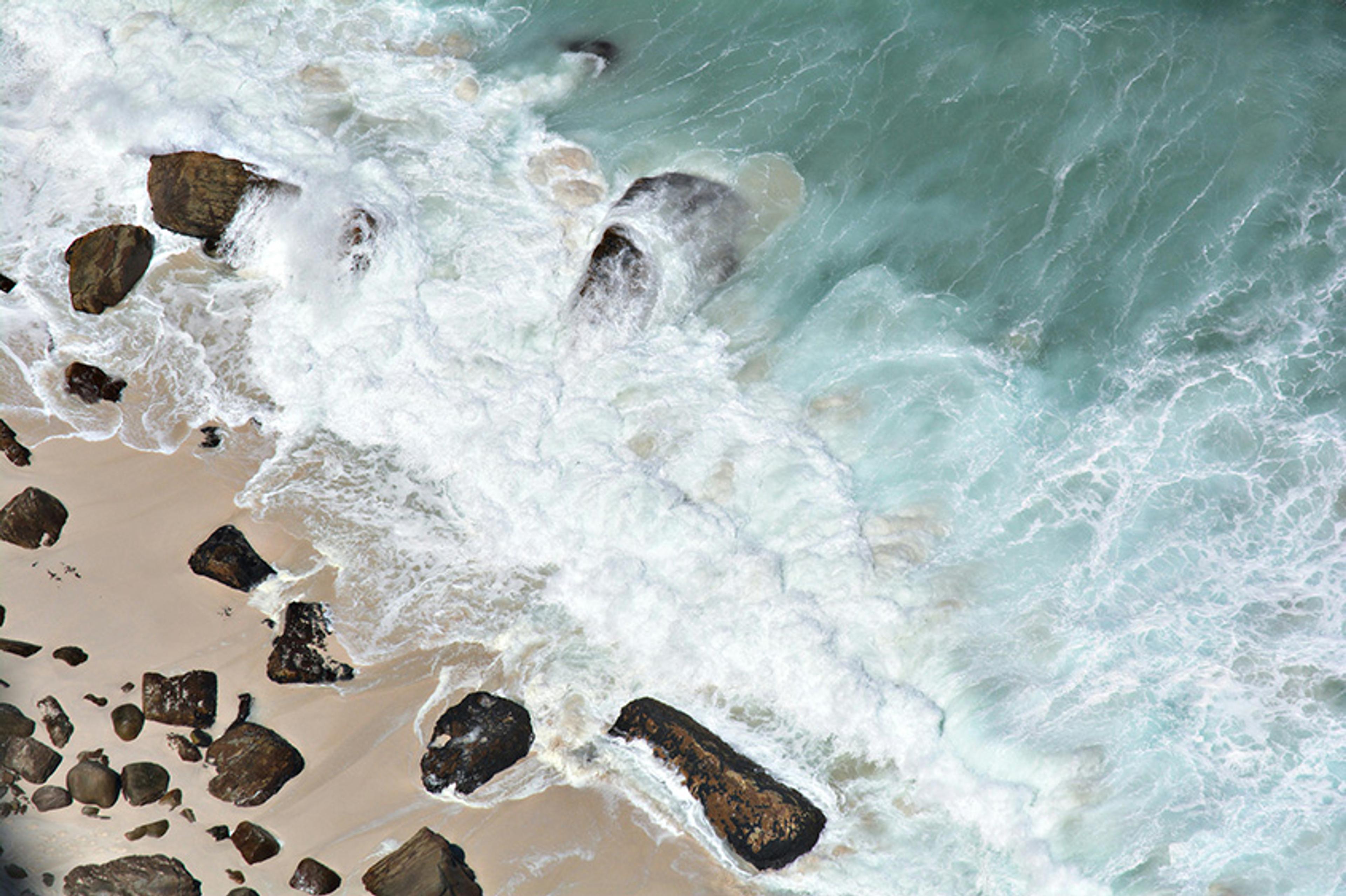 Aerial photo of turquoise waves crashing onto a sandy beach with scattered rocks.