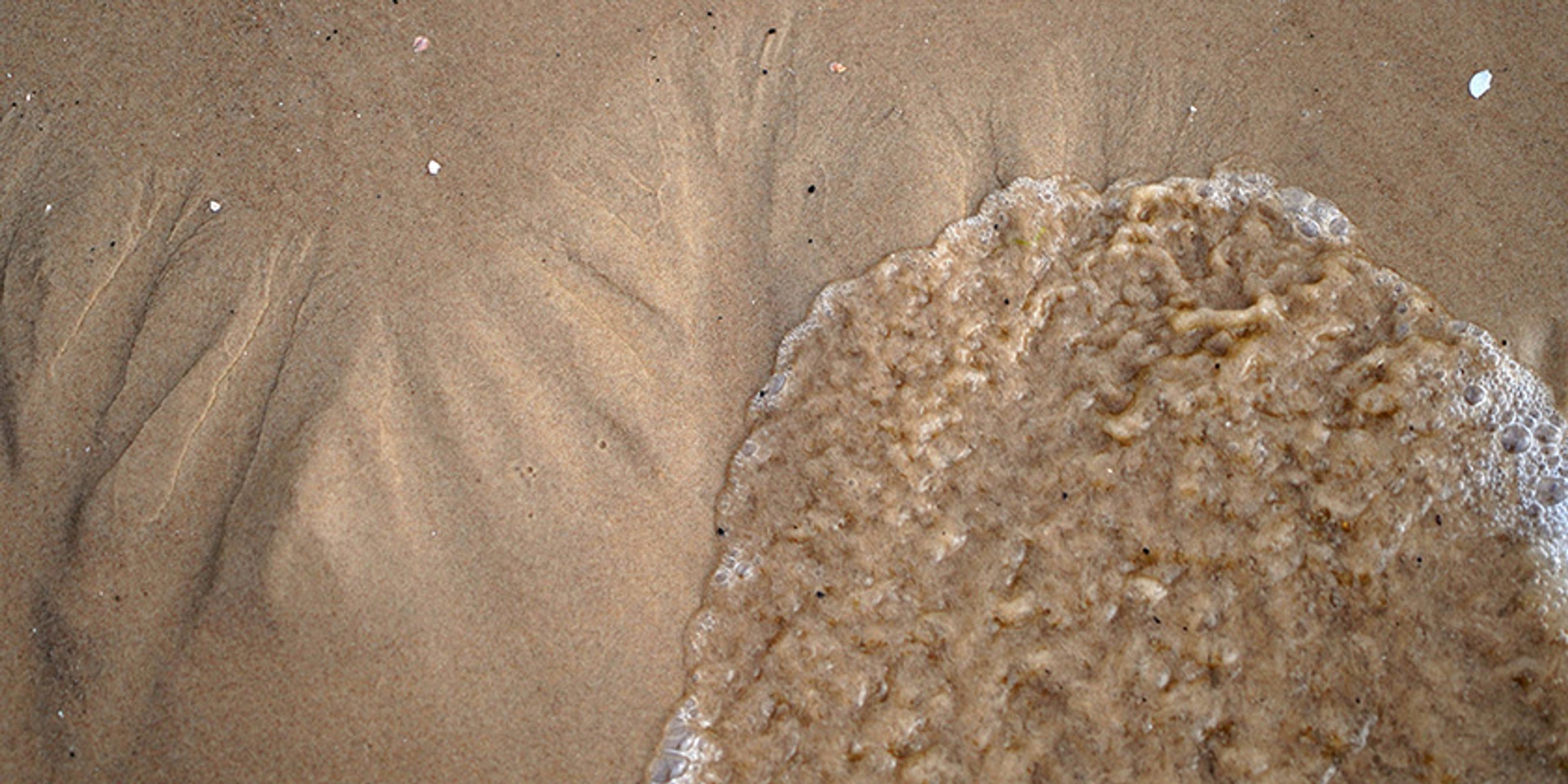 Photo of a sandy beach with small wave patterns forming ridges in the sand and water washing over part of the sand.