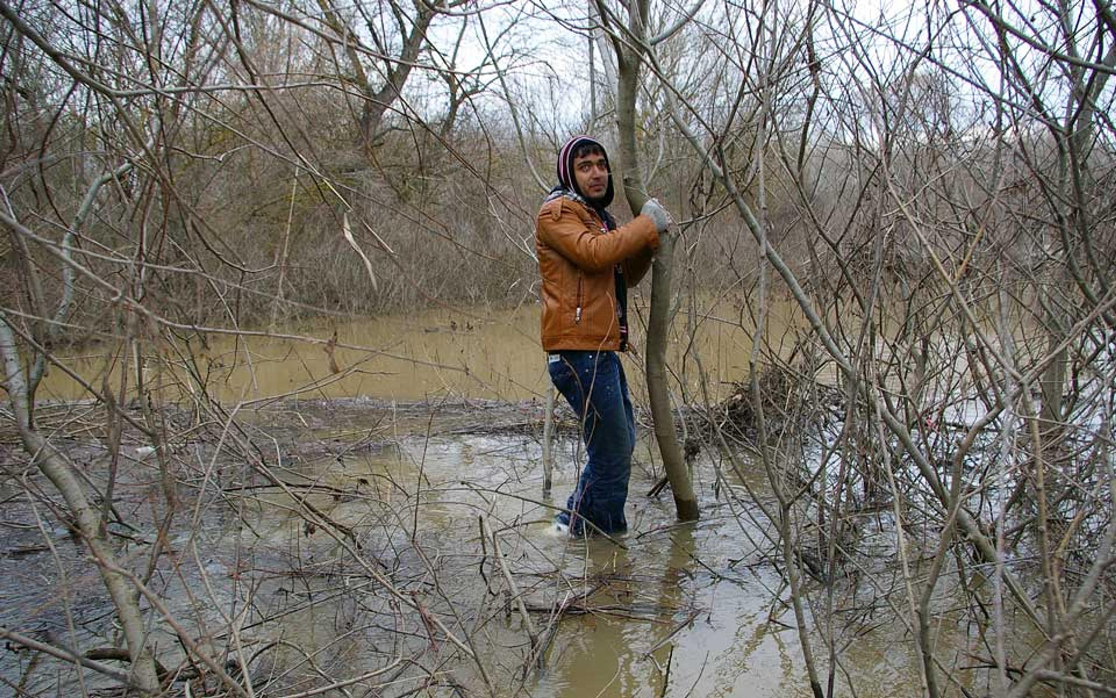 Photo of a person standing in a flooded forested area holding a tree, surrounded by leafless branches and muddy water.