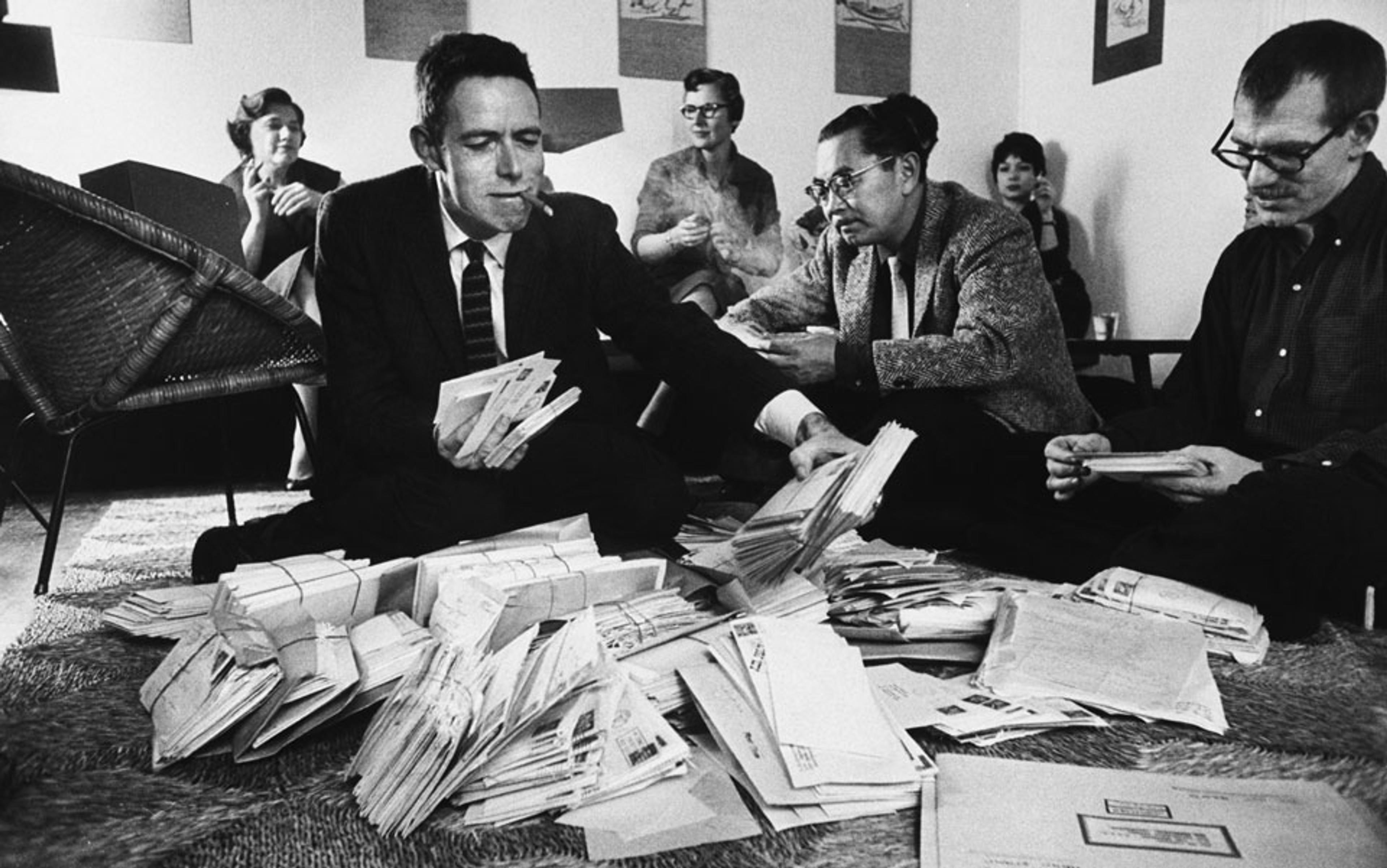 Black and white photo of a group of people sorting through a large pile of letters on the floor, some seated cross-legged, one smoking.