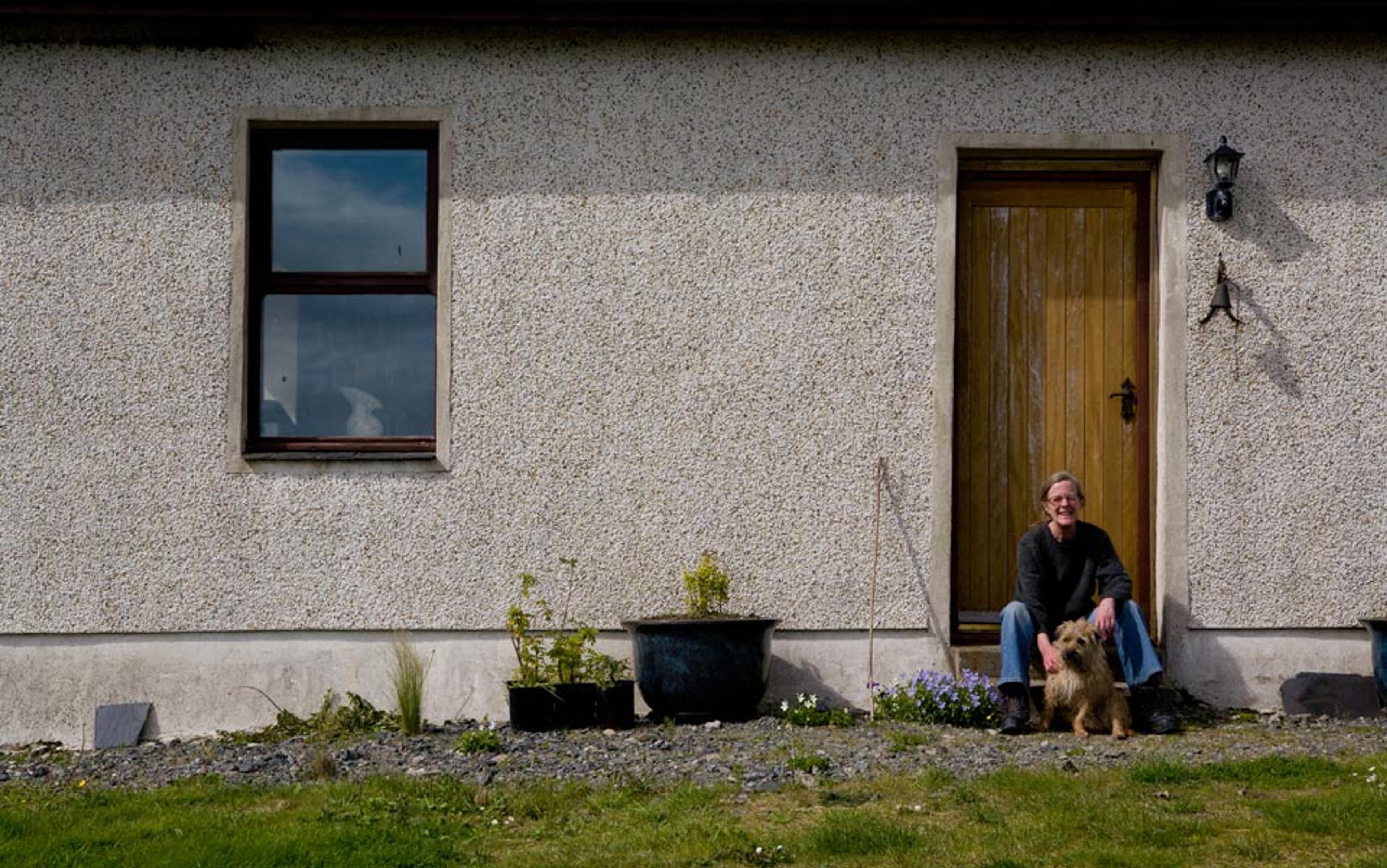 A woman sitting by a wooden door with a small dog, next to a wall with a window and some potted plants.