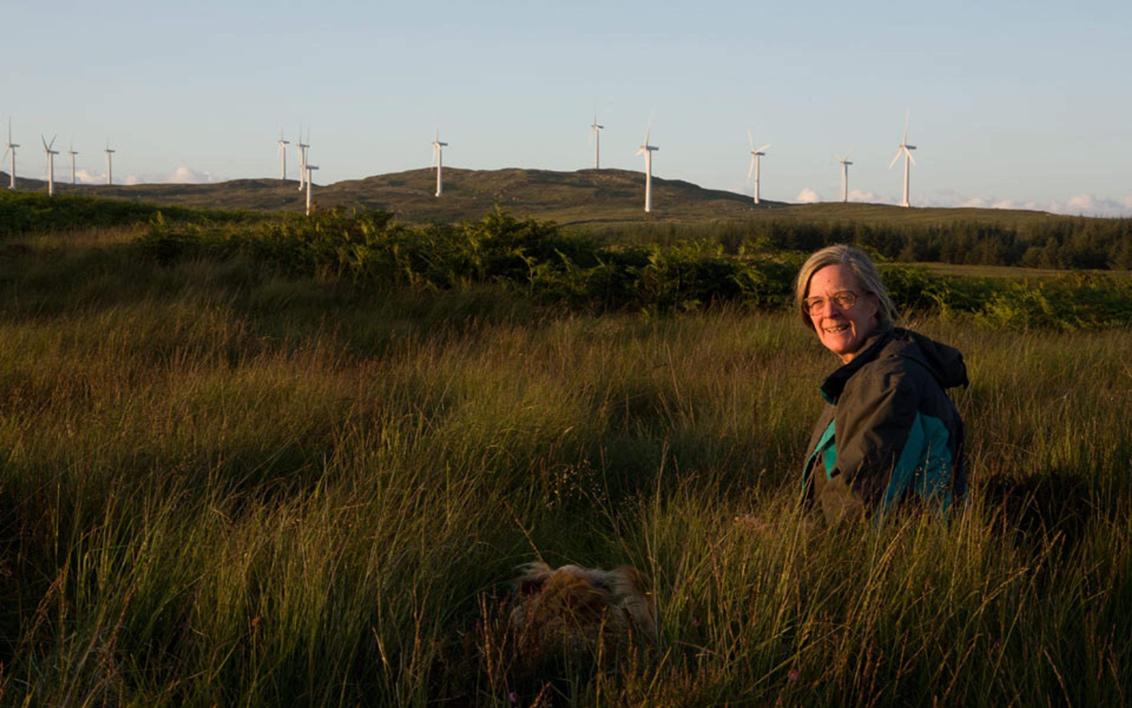 A woman in a grassy field smiling at the camera, with wind turbines on hills in the background under a clear sky.