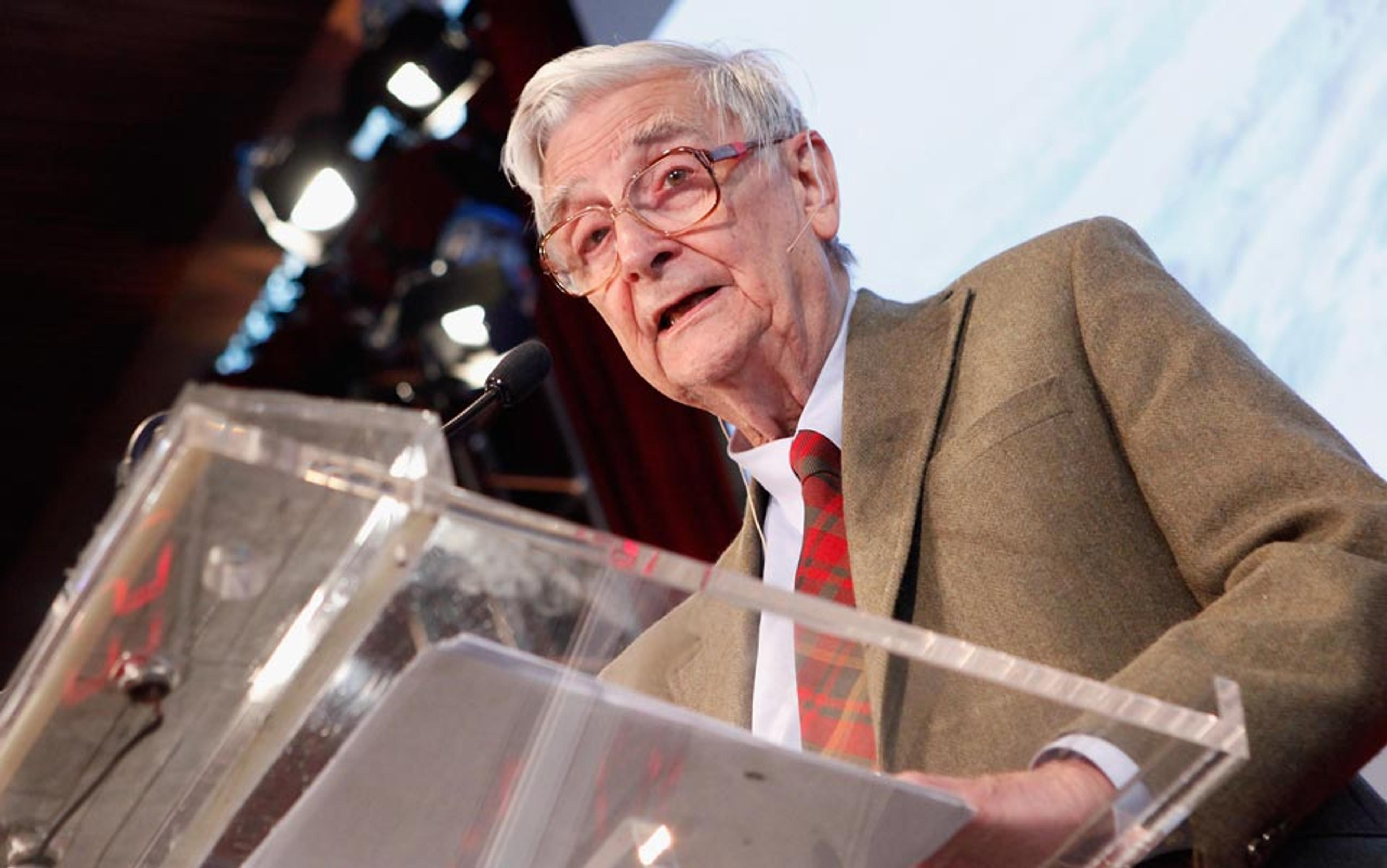 Photo of an elderly man with grey hair and glasses speaking at a podium wearing a plaid tie and suit jacket, with stage lights behind him.