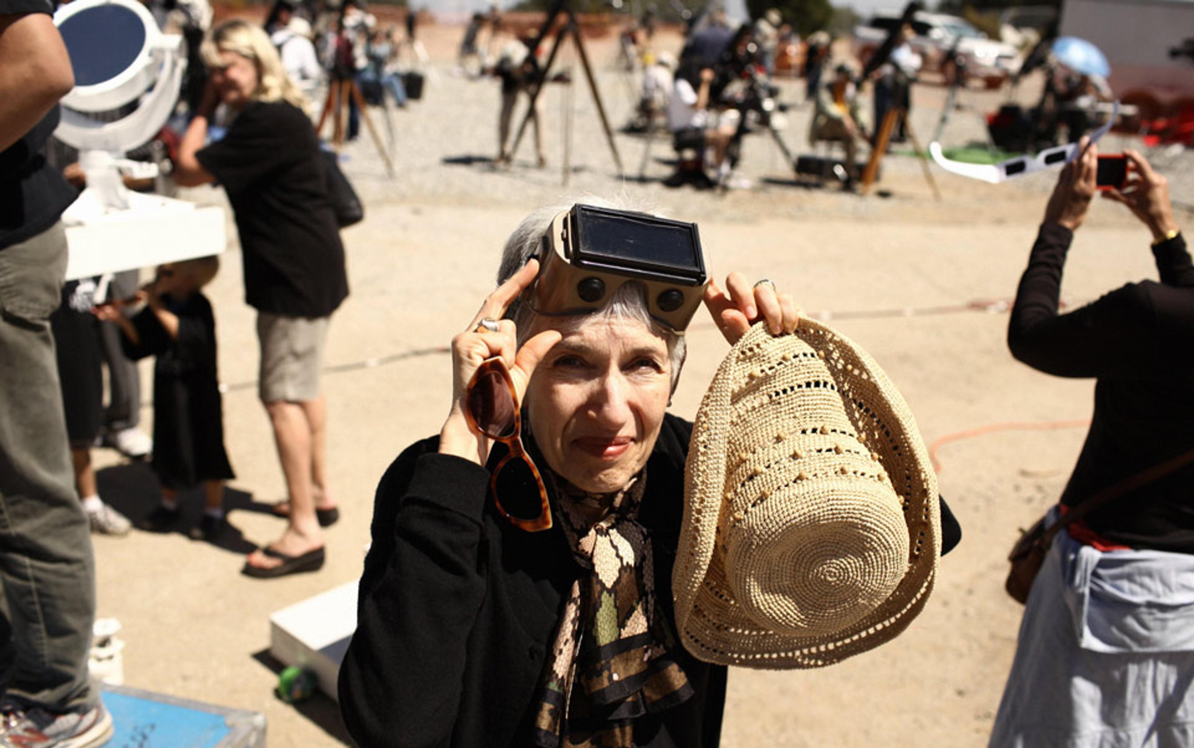 Photo of an elderly woman holding a hat, wearing solar viewing glasses outdoors with people and telescopes in the background.