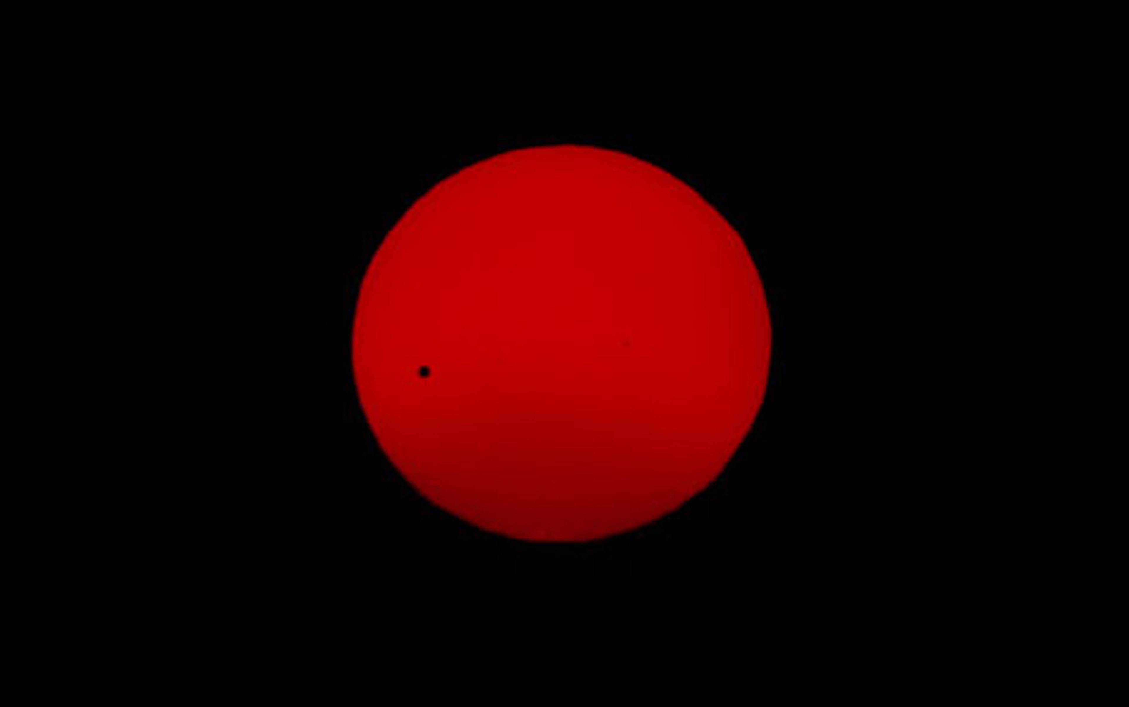 Photo of a bright red circle resembling the Sun against a black background with a tiny black dot on its surface.