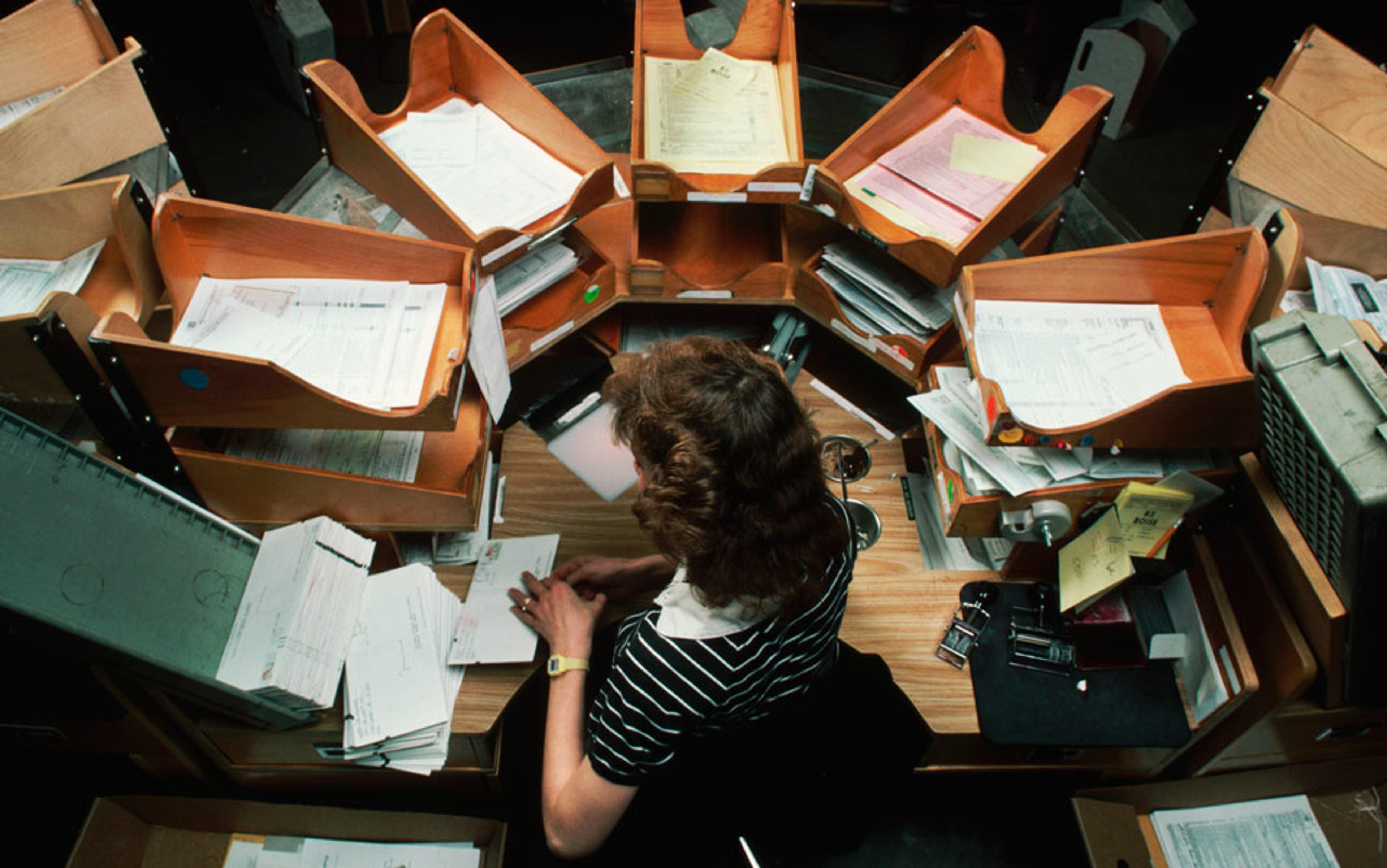 Photo of a person working at a desk surrounded by stacks of papers and wooden trays in an organised, circular arrangement.