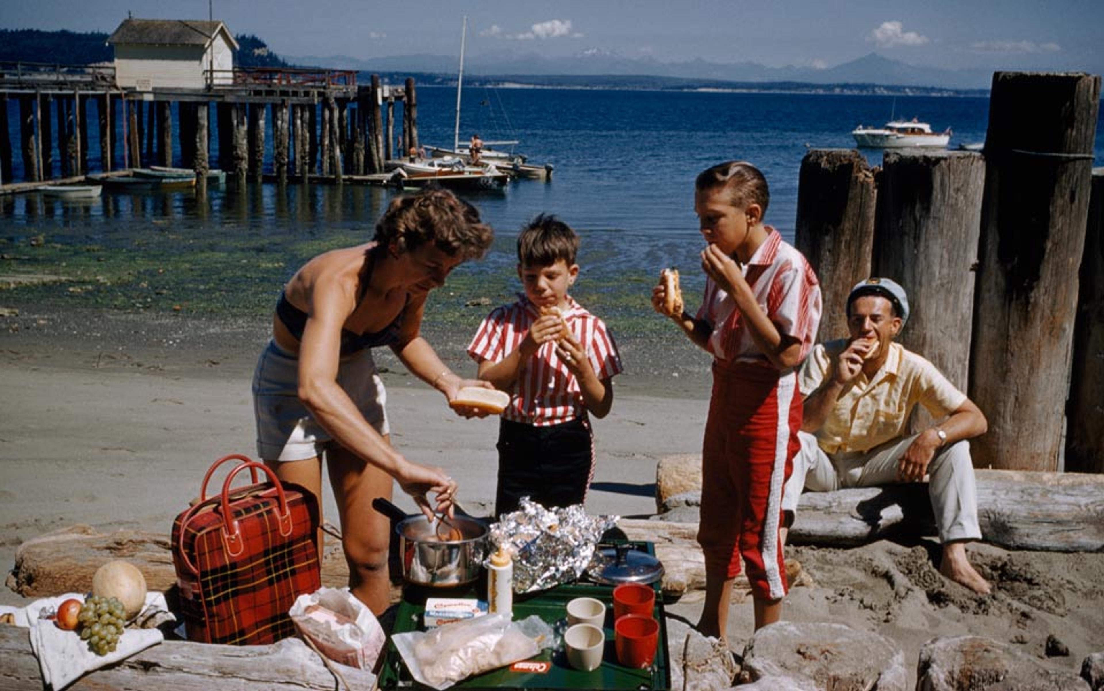 A family has a picnic on a beach with a pier in the background. Seen in a photo with the sea and a boat in the distance.