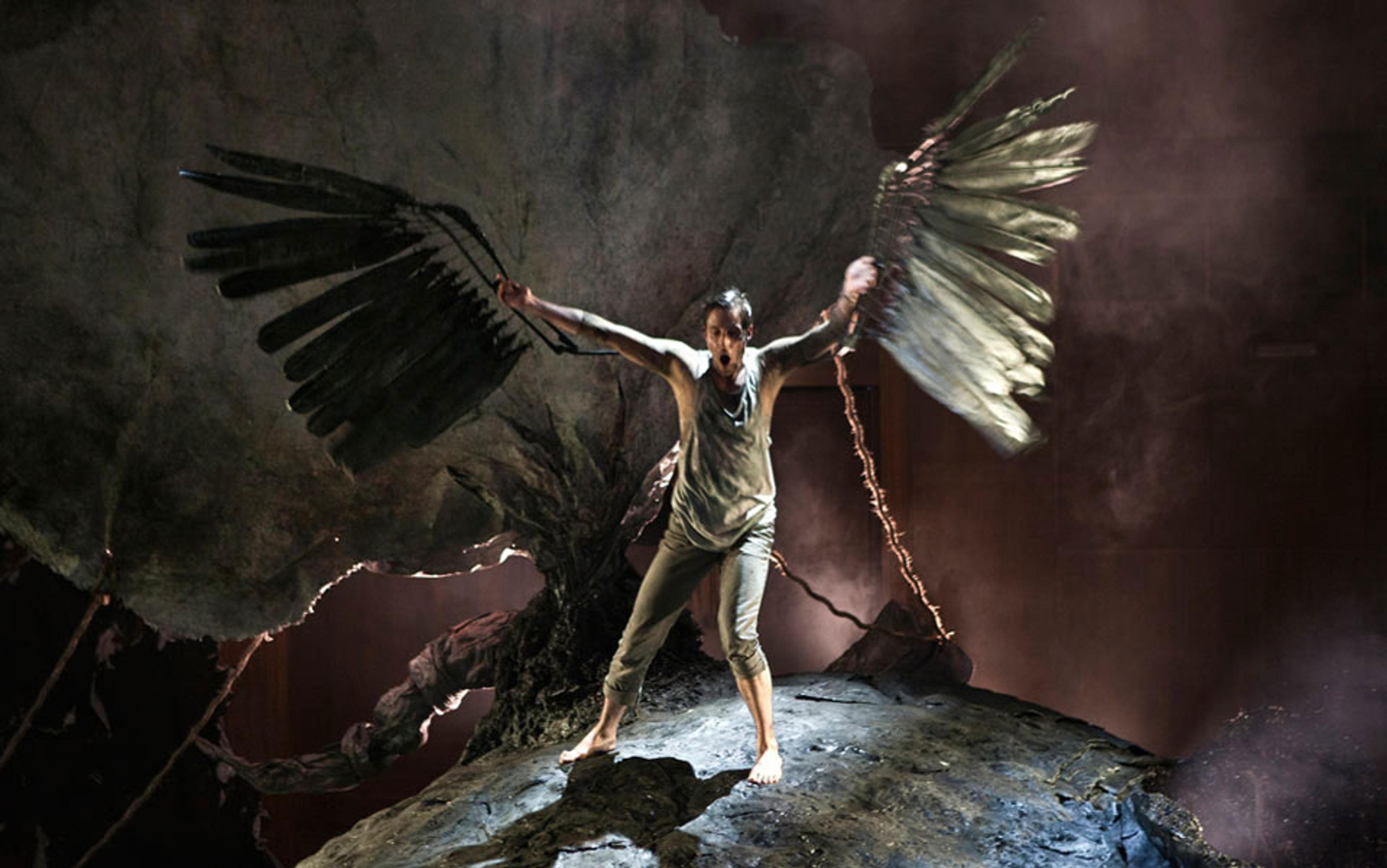 A person standing on a rock with large, dark wings in a dramatic, smoky setting, presenting an intense expression.