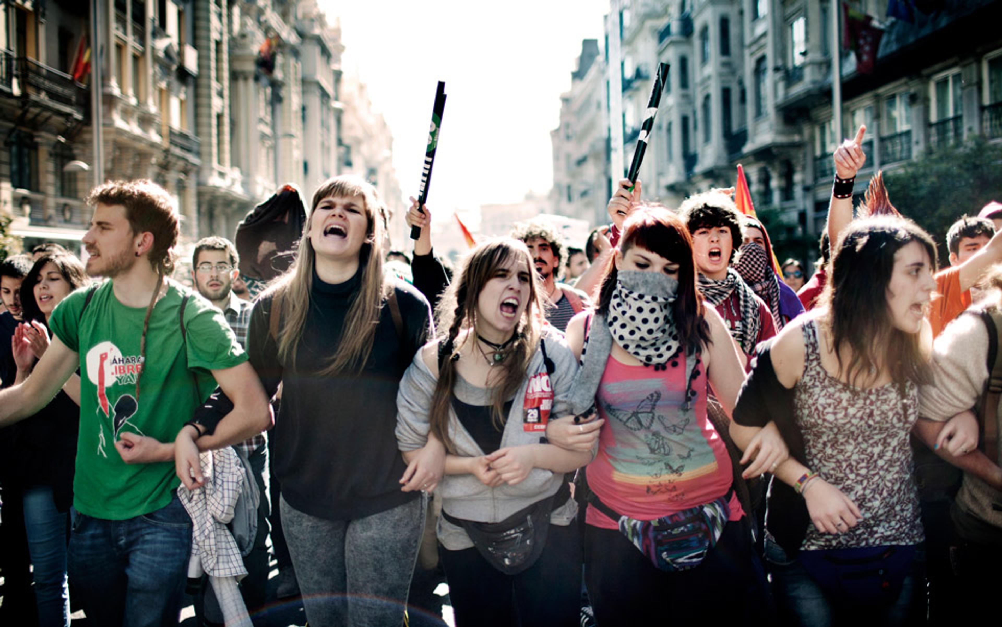 A crowd of young people protesting in a city street, shouting and holding signs while linking arms in solidarity.