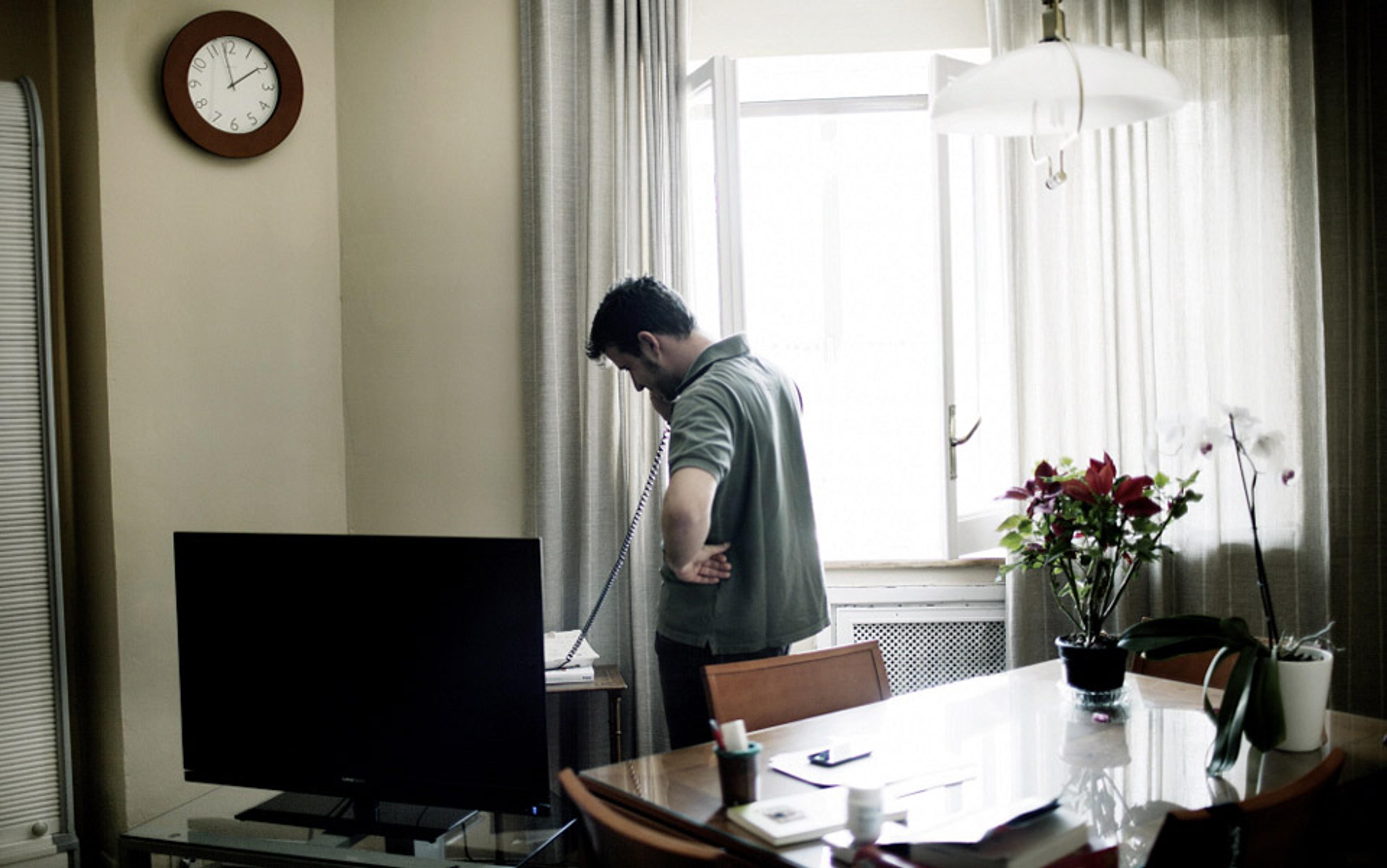 A man in a room standing by a window and talking on a landline phone, next to a dining table with flowers and a TV.